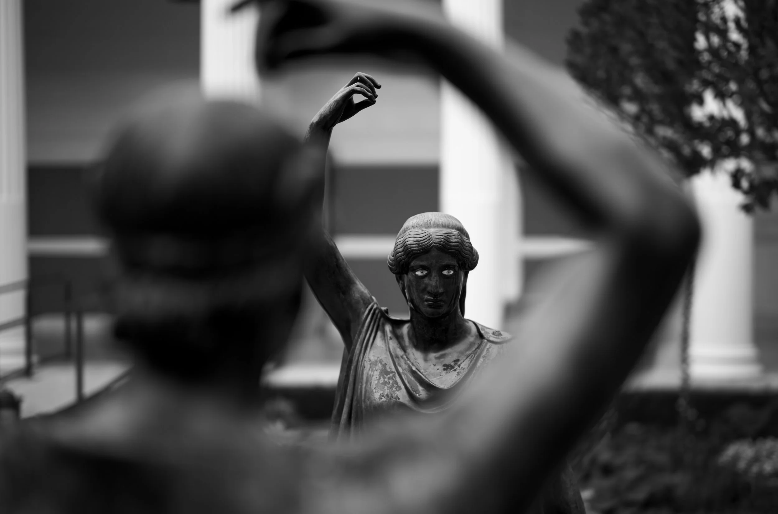 Black and white photograph of a classical sculpture of a woman, seen through the opening of another sculpture, with a blurred background including trees and a building. Getty Villa, Pacific Palisades, CA.