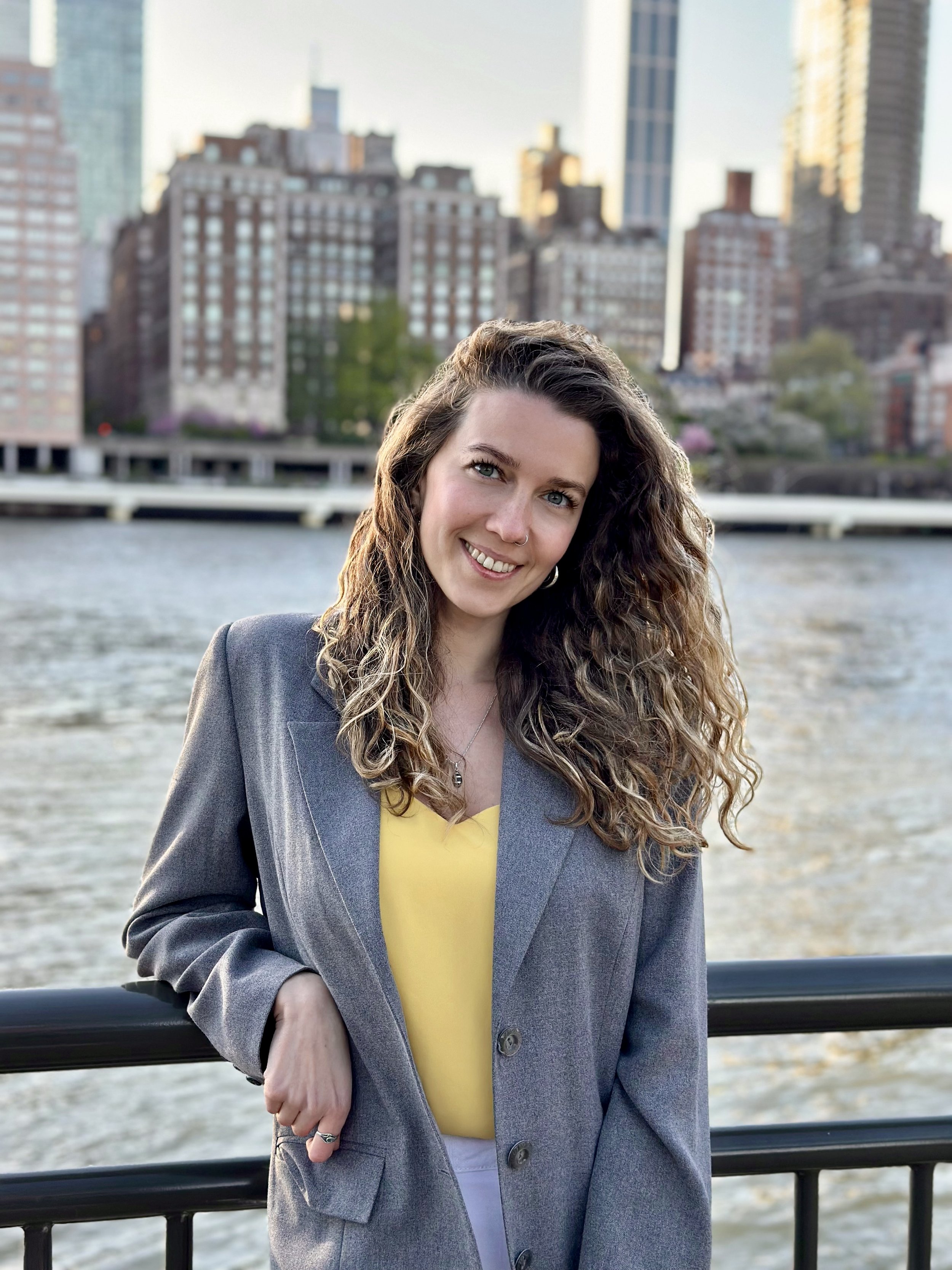 A woman with long curly hair, wearing a gray blazer over a yellow top, standing outdoors by a body of water with city buildings in the background.