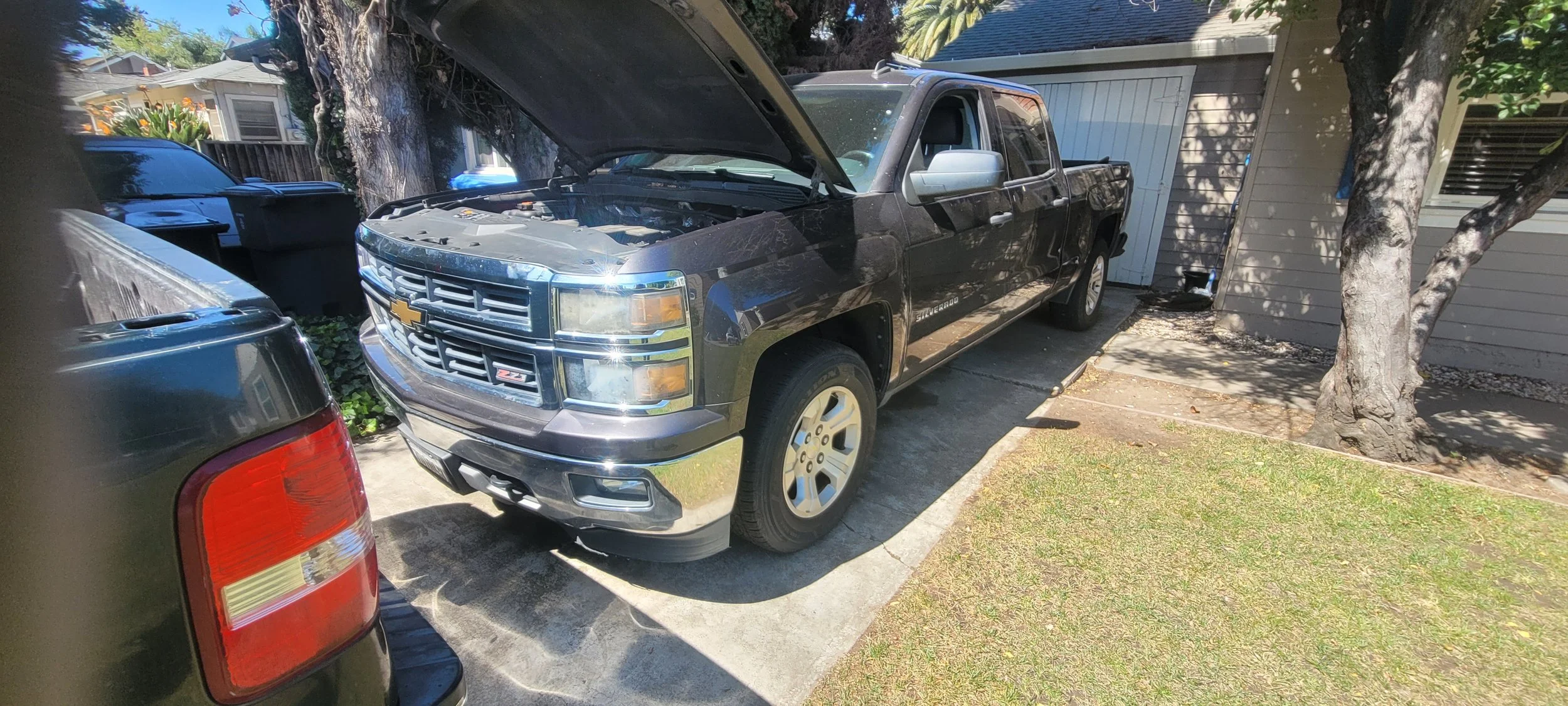 Black Chevrolet Silverado pickup truck with open hood parked in driveway next to a tree and a shed.