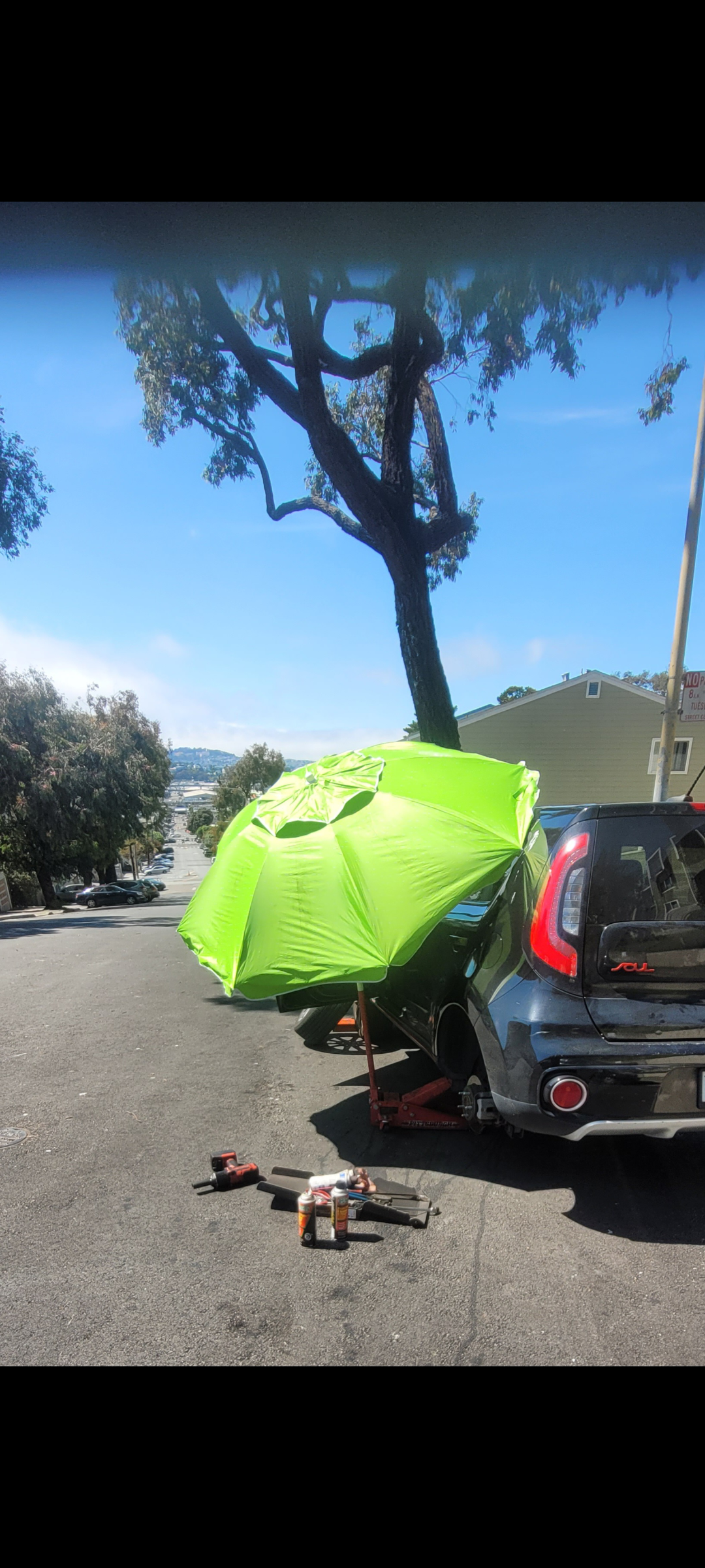A parking lot with a black SUV that has a green umbrella attached to it, tools, and spray cans on the ground nearby. A tall tree and buildings are visible in the background.