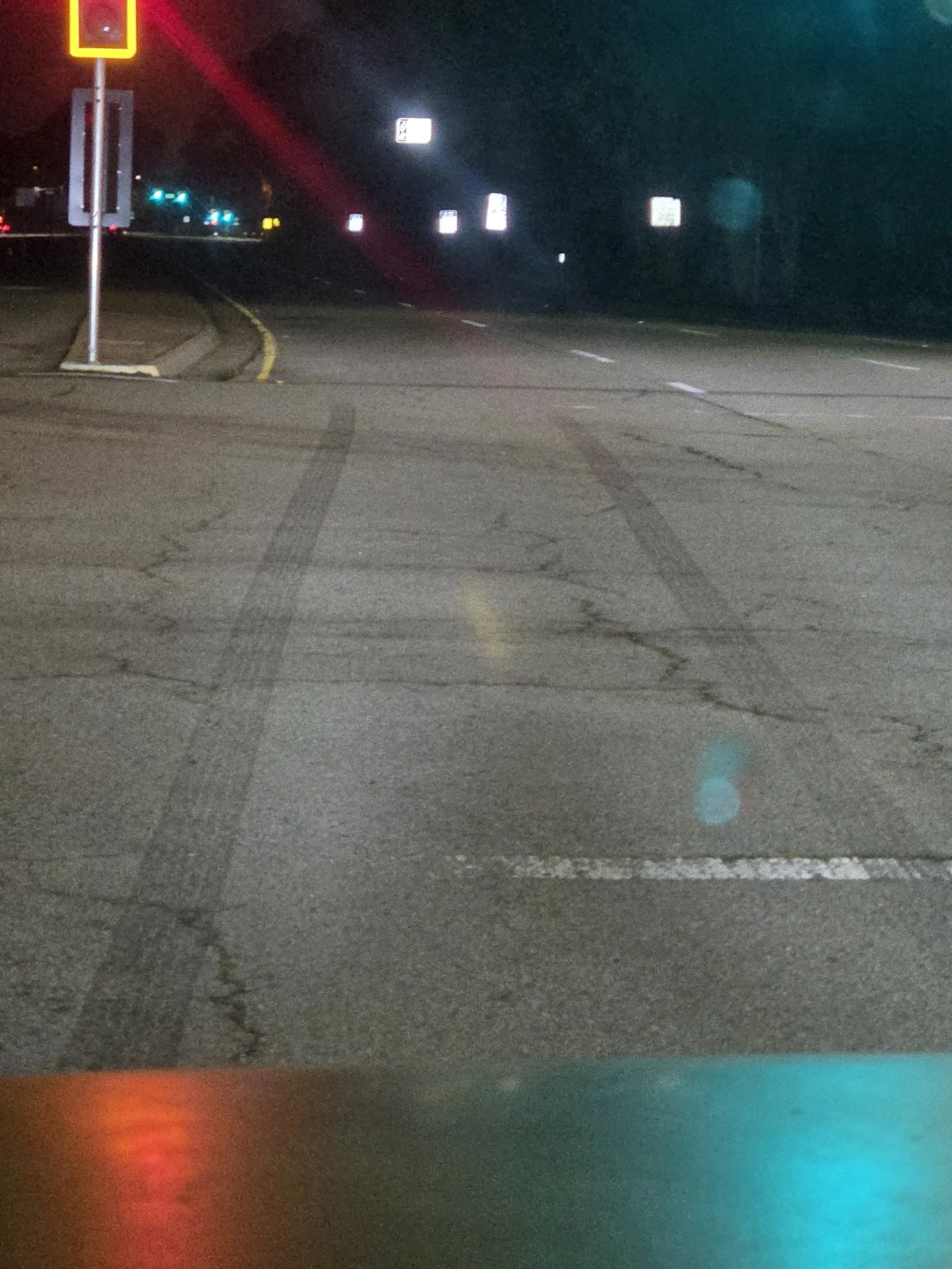 Nighttime view of an empty intersection with streetlights and traffic signs, with visible tire marks on the cracked asphalt road.