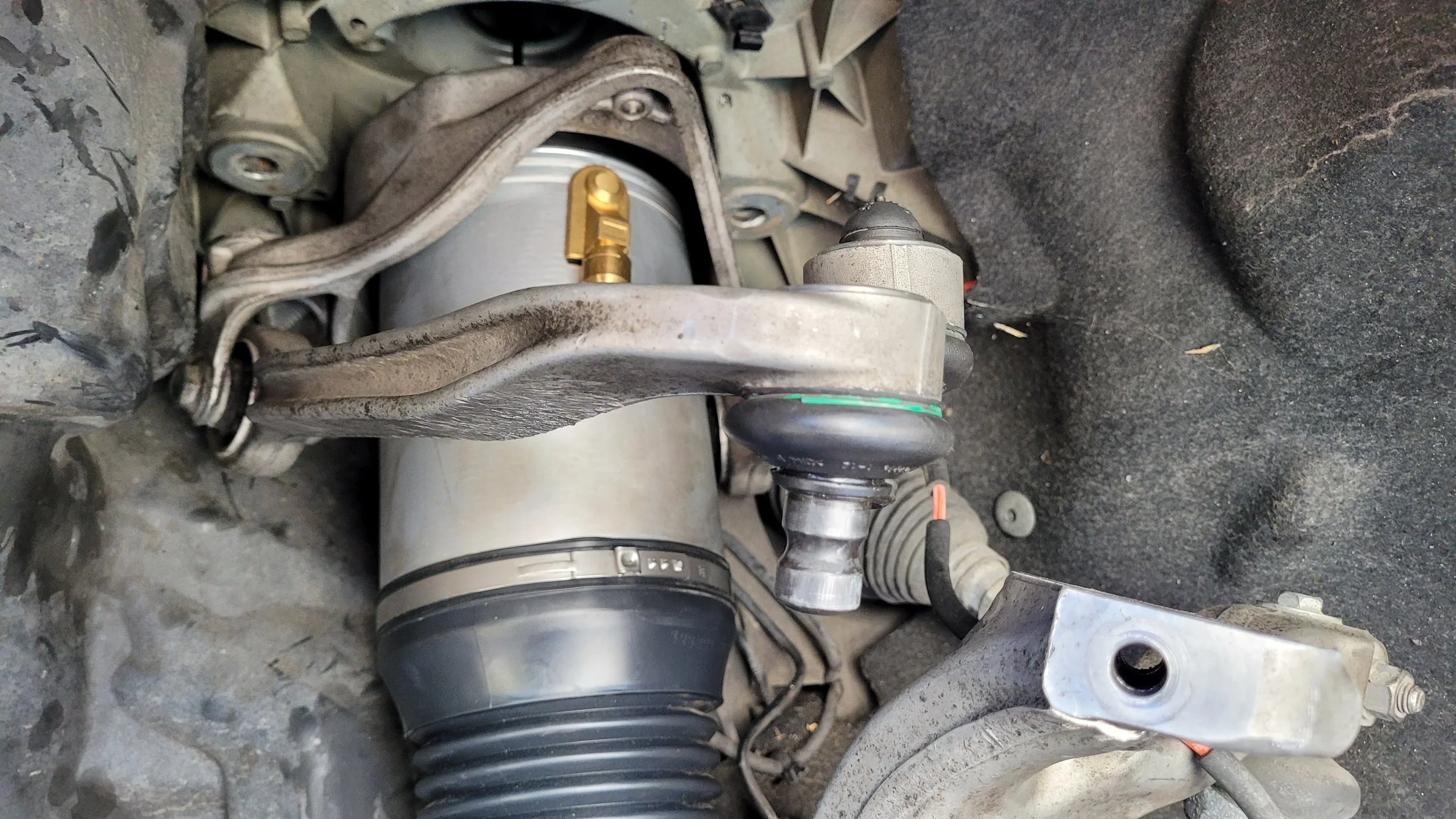 Close-up view of automotive suspension and steering components, including a strut, control arm, and various bolts and joints, on a vehicle in a garage or workshop setting.