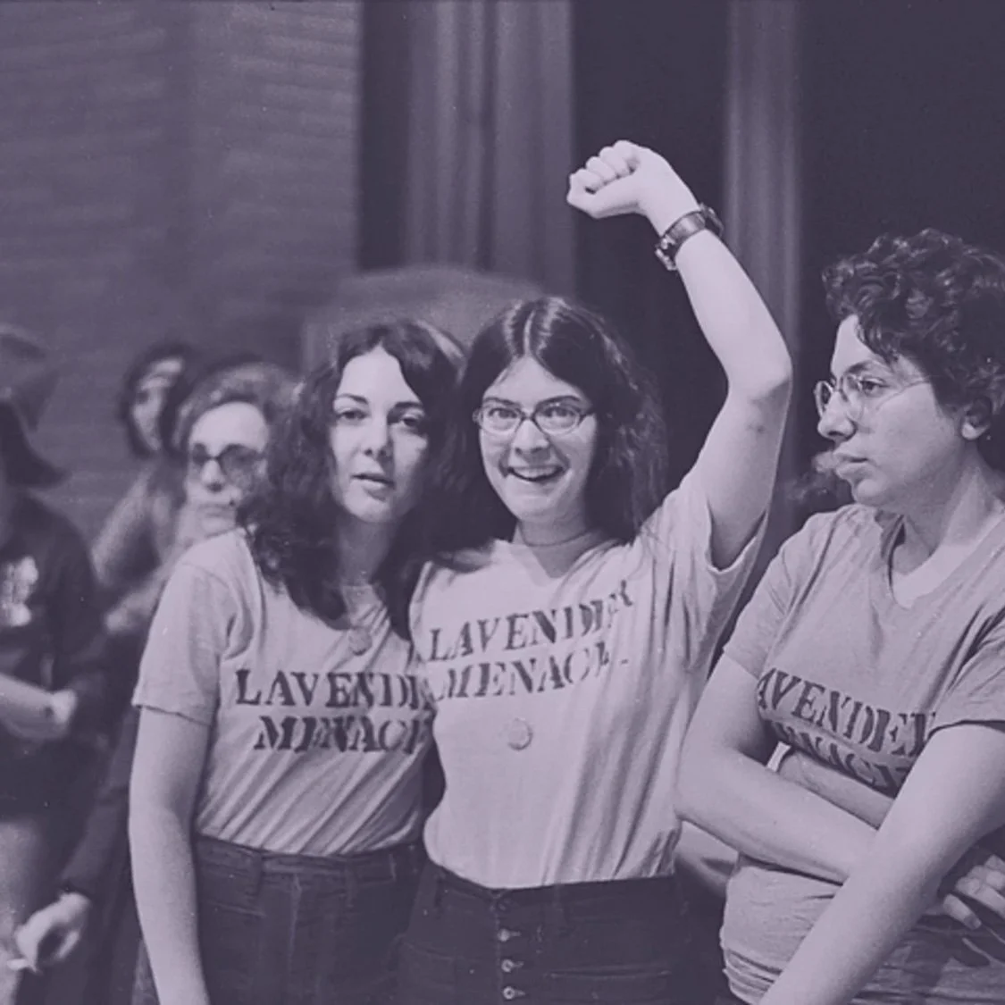 Black and white photo of three women wearing matching t-shirts that say 'Lavender Menace.' The woman in the middle is smiling and raising her arm, while the other two look on.