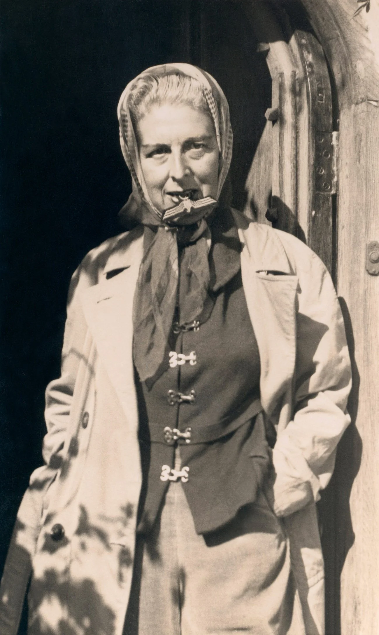 Black and white photograph of an elderly woman wearing a headscarf, a light-colored coat, and a dark dress with toggle buttons. She is standing outdoors next to a wooden door or wall, smiling slightly with her hands in her pockets.