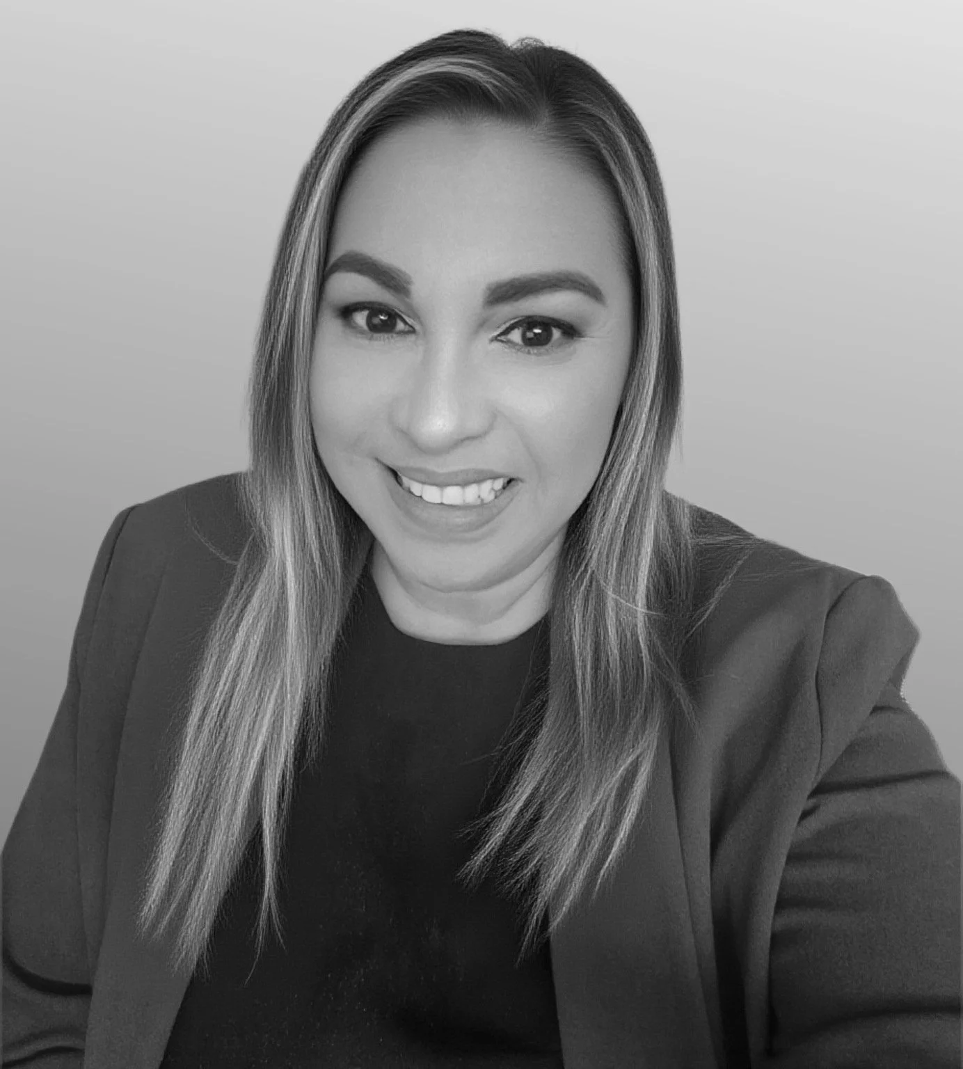 A woman with long dark hair, smiling and sitting with arms crossed, wearing a blazer and white shirt in a grayscale photo.