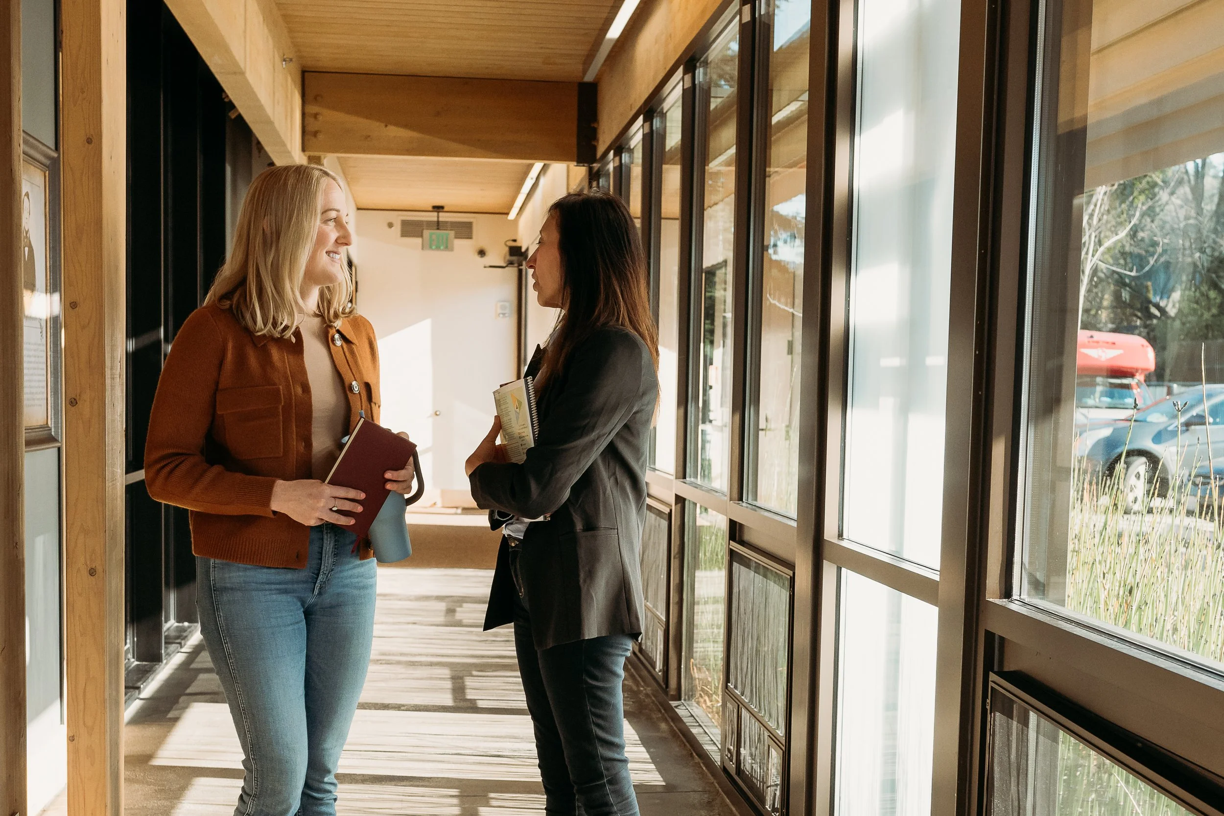 two women standing in a sunlit hallway facing each other and conversing