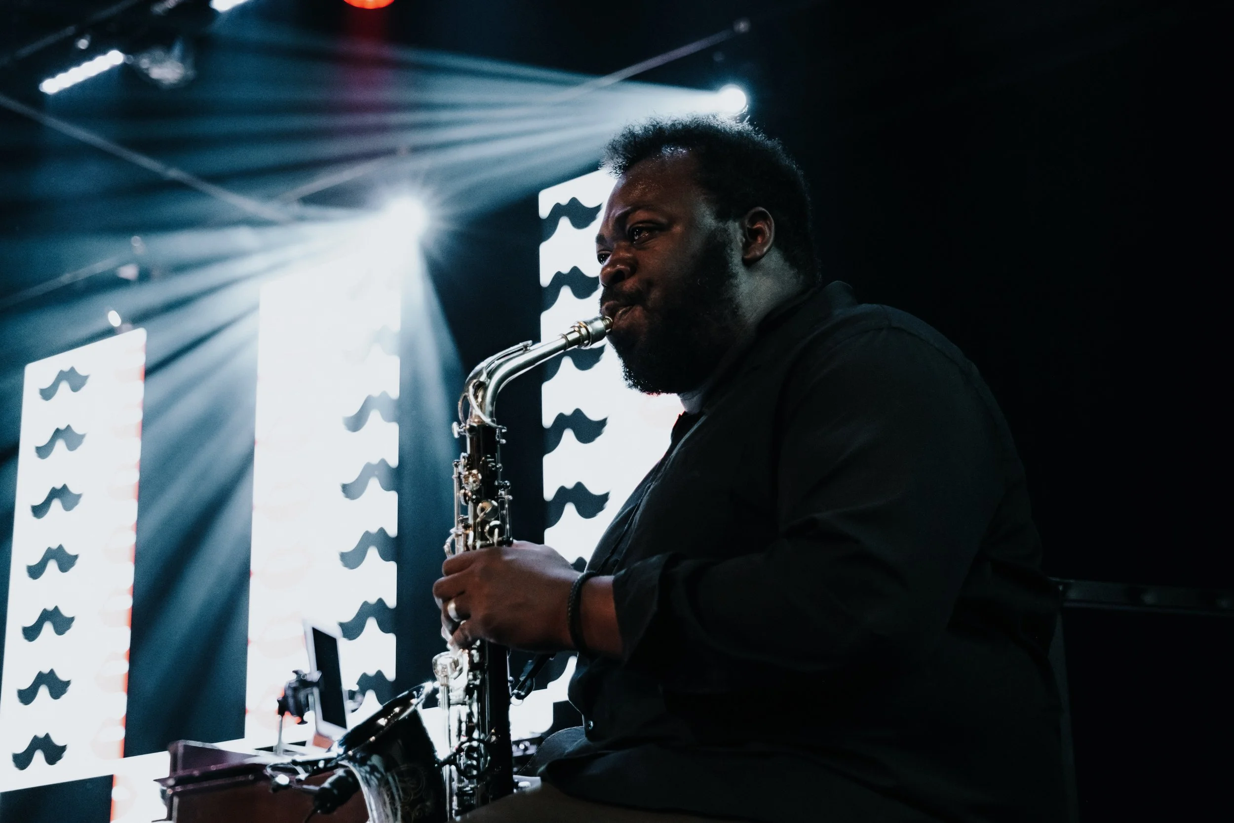 A man playing the saxophone on stage with bright lights and patterned background.