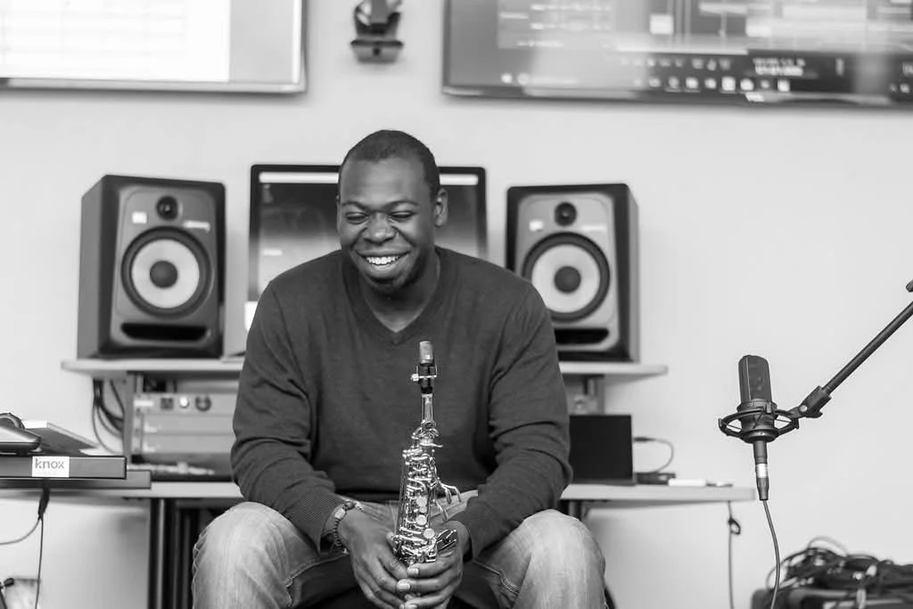 A man sitting in a music studio, holding a saxophone, and smiling. There are studio monitors, a microphone, and audio equipment in the background.
