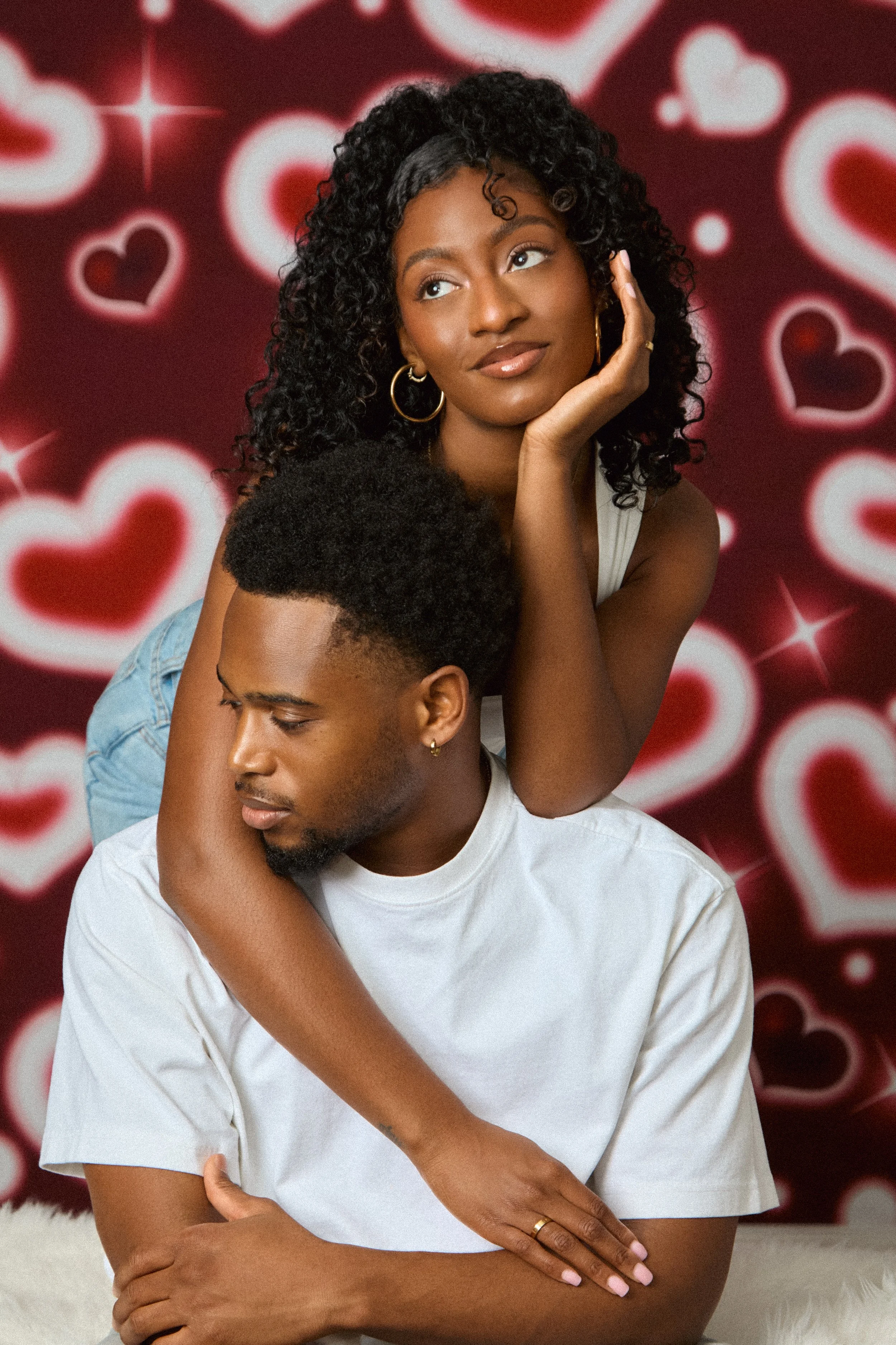 A young couple sitting together, the woman with curly hair resting her face on her hand and looking into the distance, while the man has his eyes closed and is leaning into her shoulder with his arms crossed. The background features a red backdrop with white and red heart patterns.