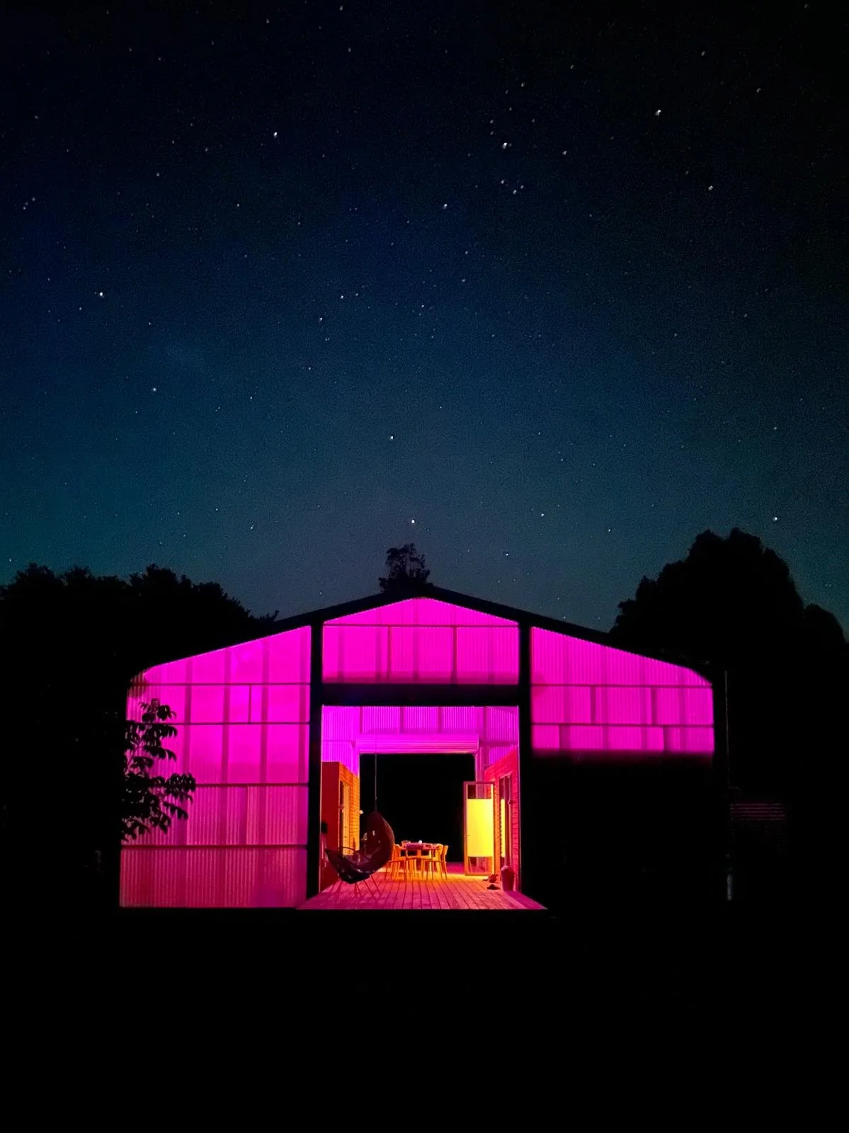 A modern home using translucent walls backlit with pink light, set against a starry night sky with trees in a garden designed by Mark Newdick in the background.