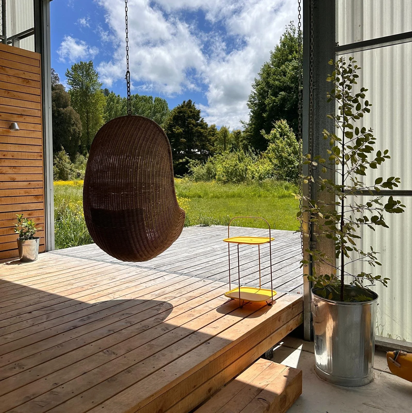 View of a wooden deck with hanging wicker chair, small yellow side table, potted plant, and lush green trees in a bright outdoor setting under a blue sky with clouds.