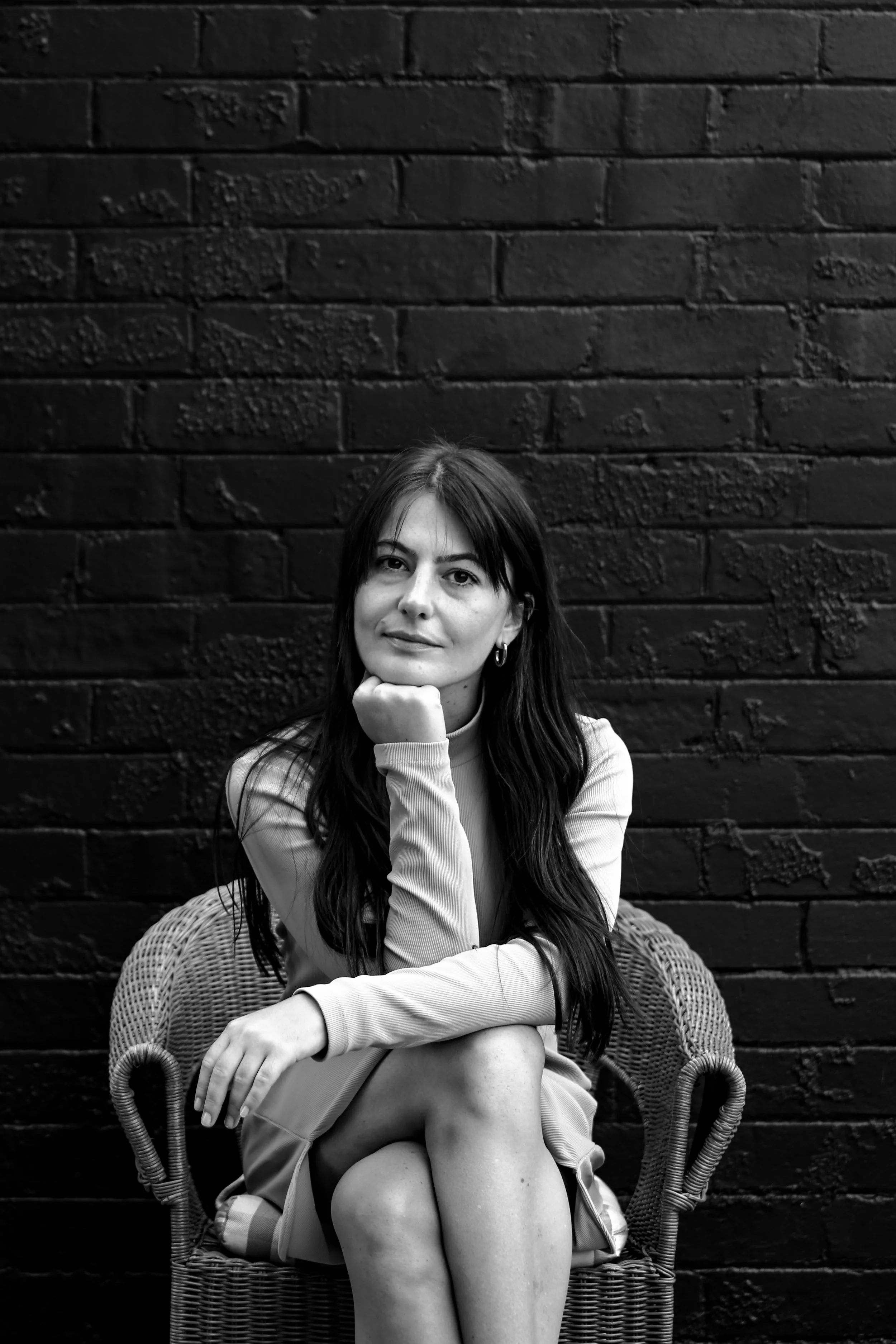 Black and white photo of a woman sitting on a wicker chair in front of a brick wall, resting her chin on her hand, wearing a long-sleeve top and a skirt.