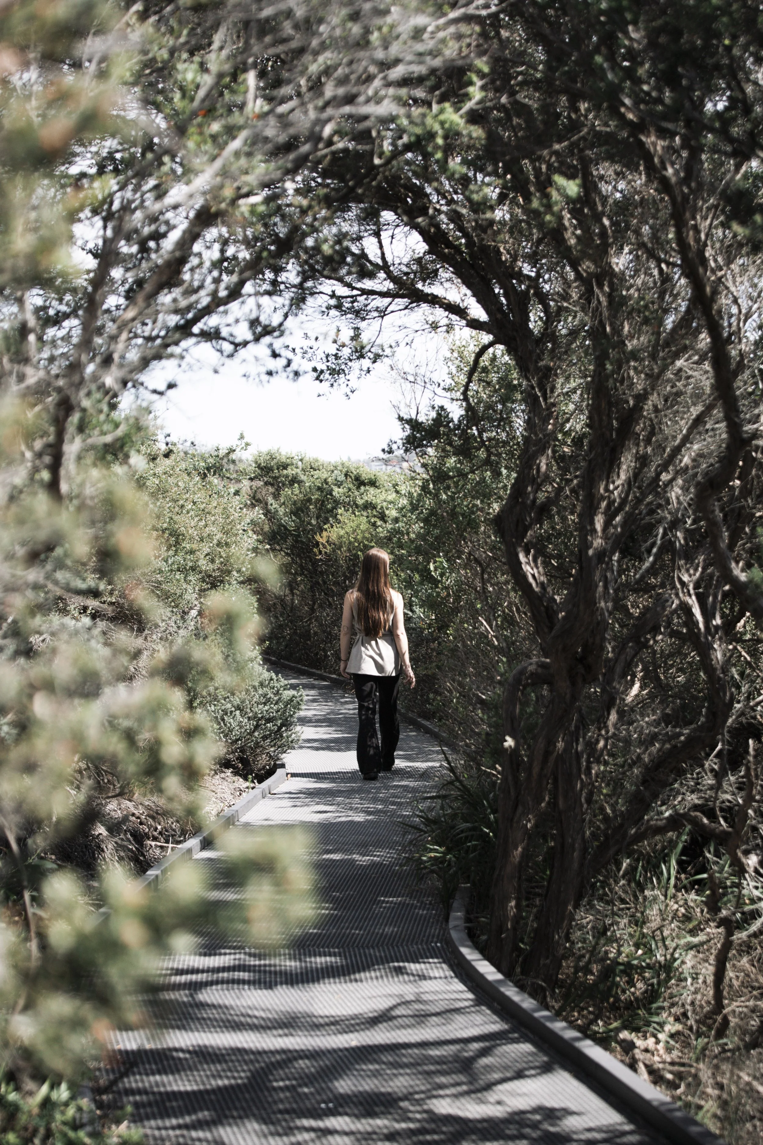A woman walking on a winding metal path through a wooded area with dense green foliage and twisted tree branches.