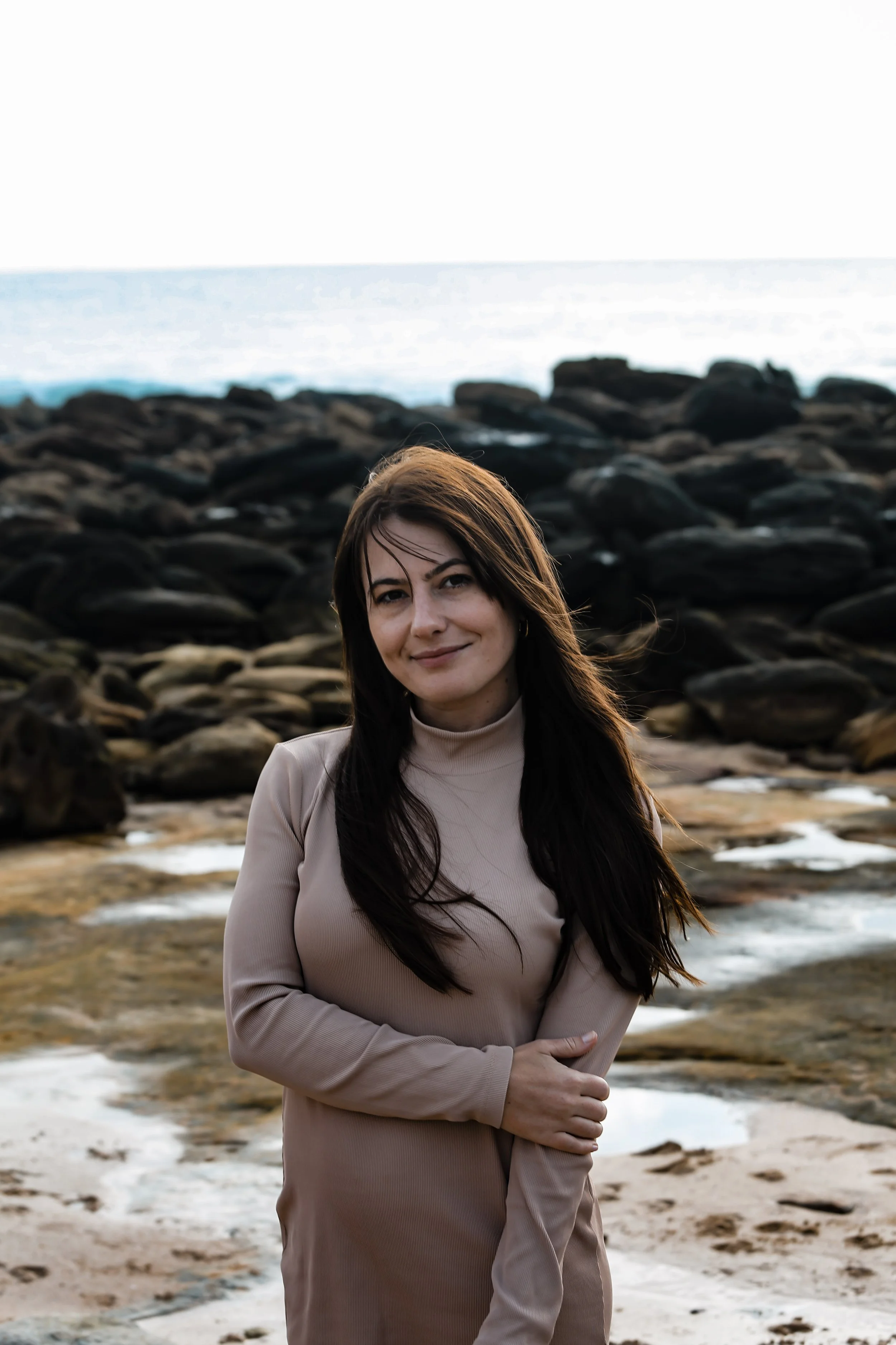 A woman with long dark hair standing on a rocky beach, smiling, with the ocean and rocks in the background.