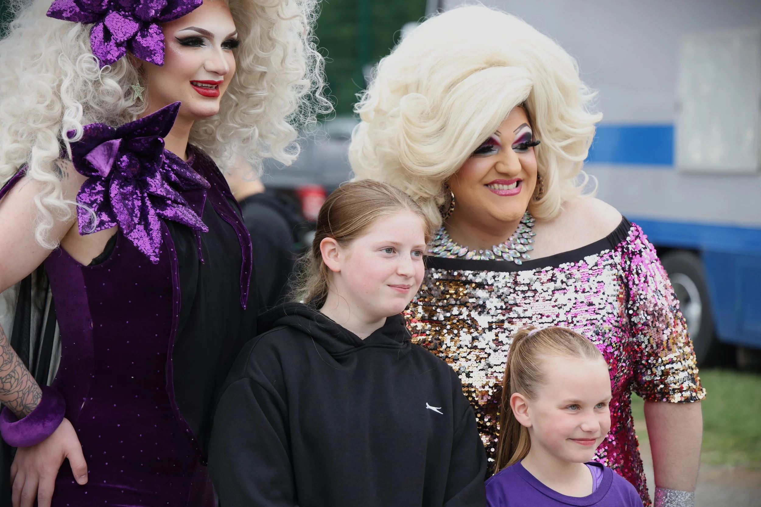Group of people, including drag queens and children, smiling outdoors.
