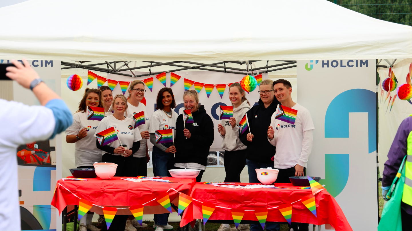 Group of smiling people holding small rainbow pride flags at an outdoor event with rainbow decorations and banners for Holcim.