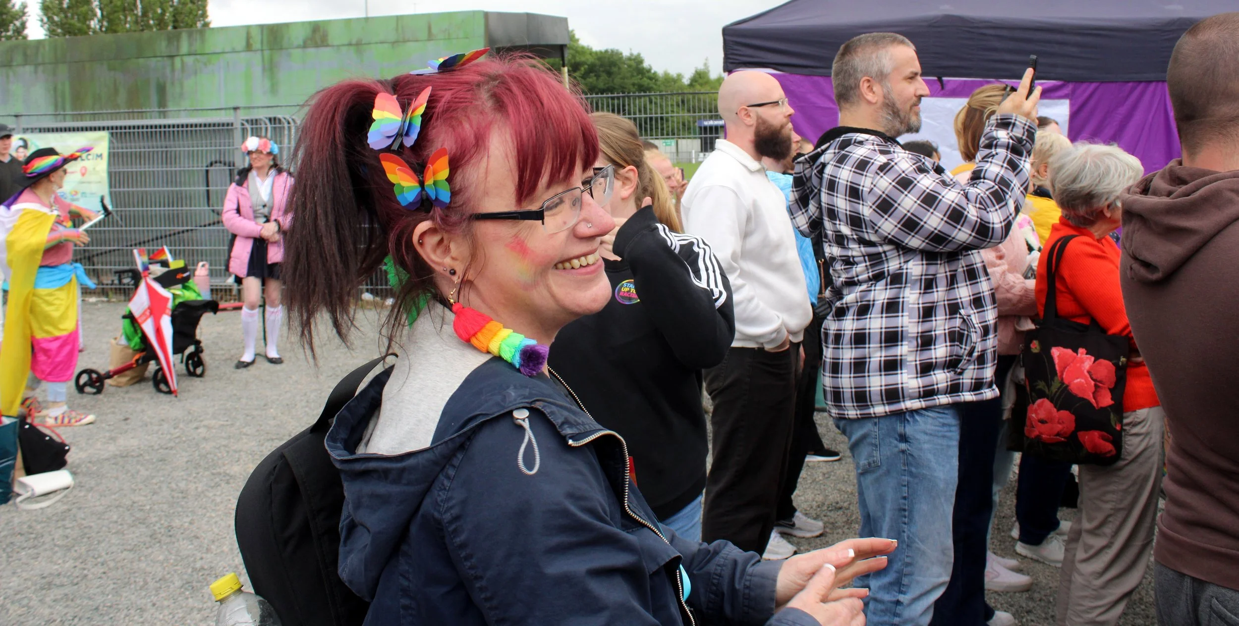 A woman with red hair and rainbow butterfly hair clips is smiling and clapping at an outdoor event. She is wearing rainbow-colored accessories and a dark jacket. Other people are standing nearby, some taking photos, at what appears to be a pride or L