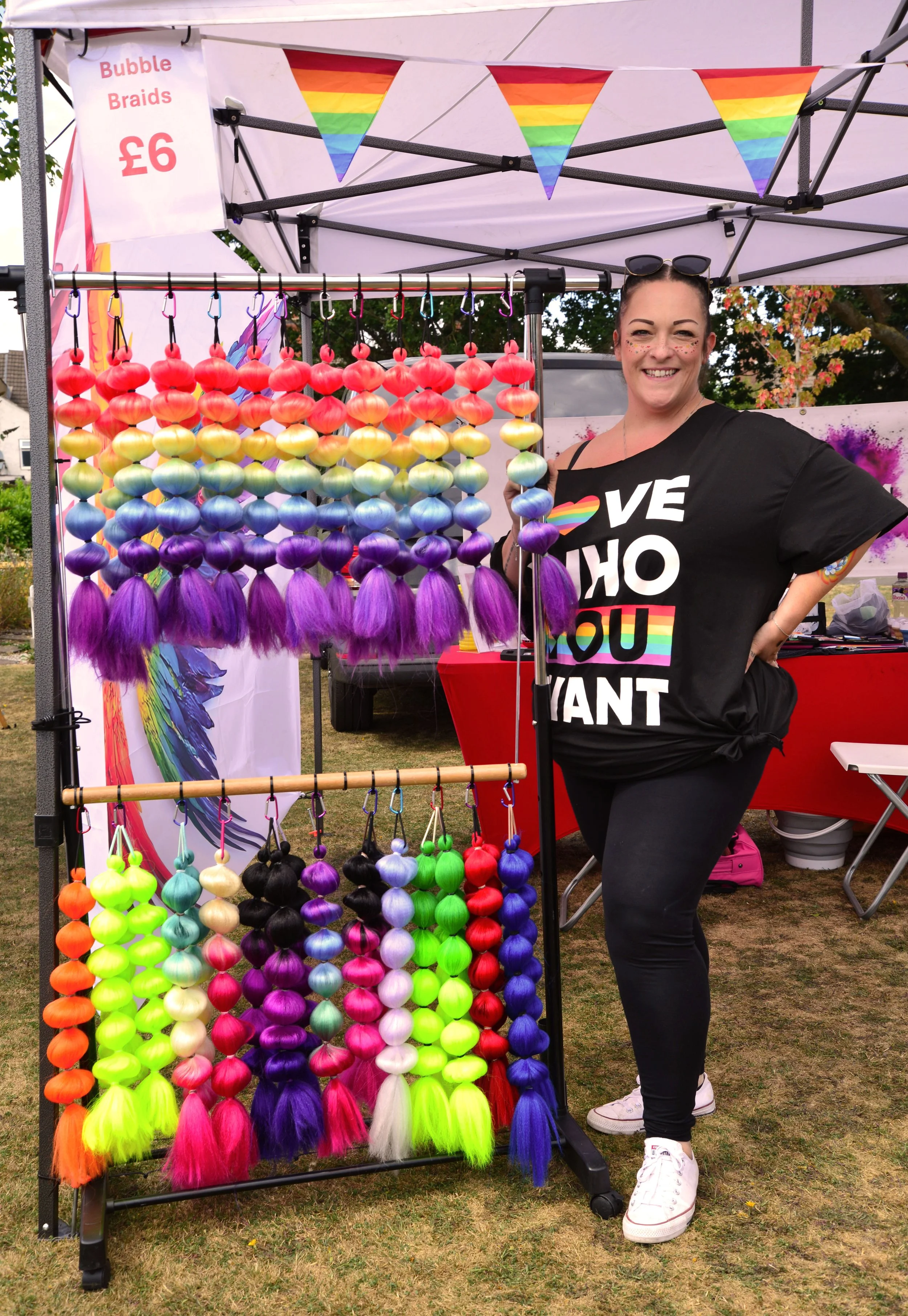 People attending a Leicester LGBTQ+ Centre event outdoors under a tent decorated with rainbow pride flags. Some individuals are gathered around a table with rainbow-colored items and balloons. A person in a wheelchair and others are engaged in activities.