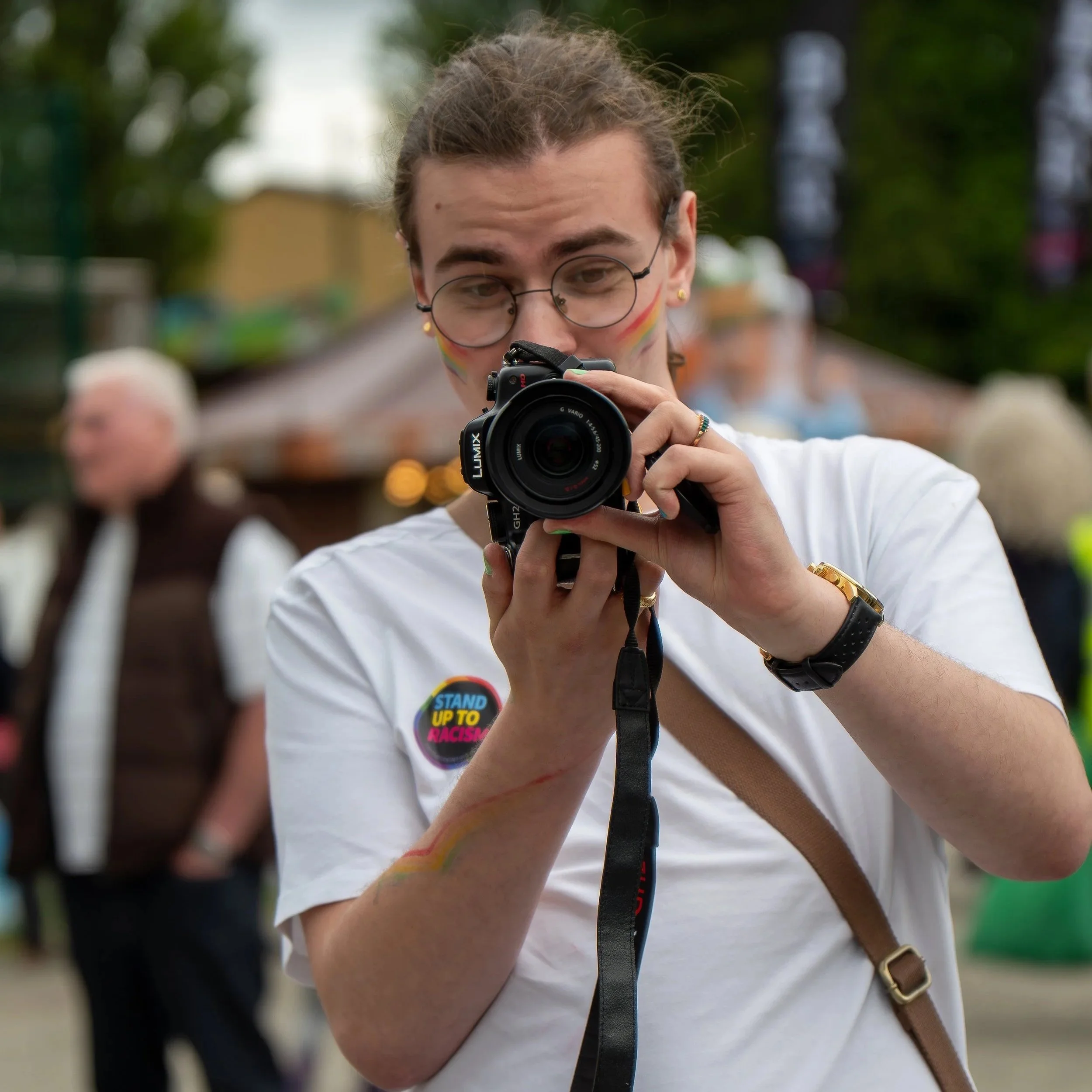Person with rainbow sticker on white shirt taking a photograph with a camera in an outdoor setting, with blurred people in the background.