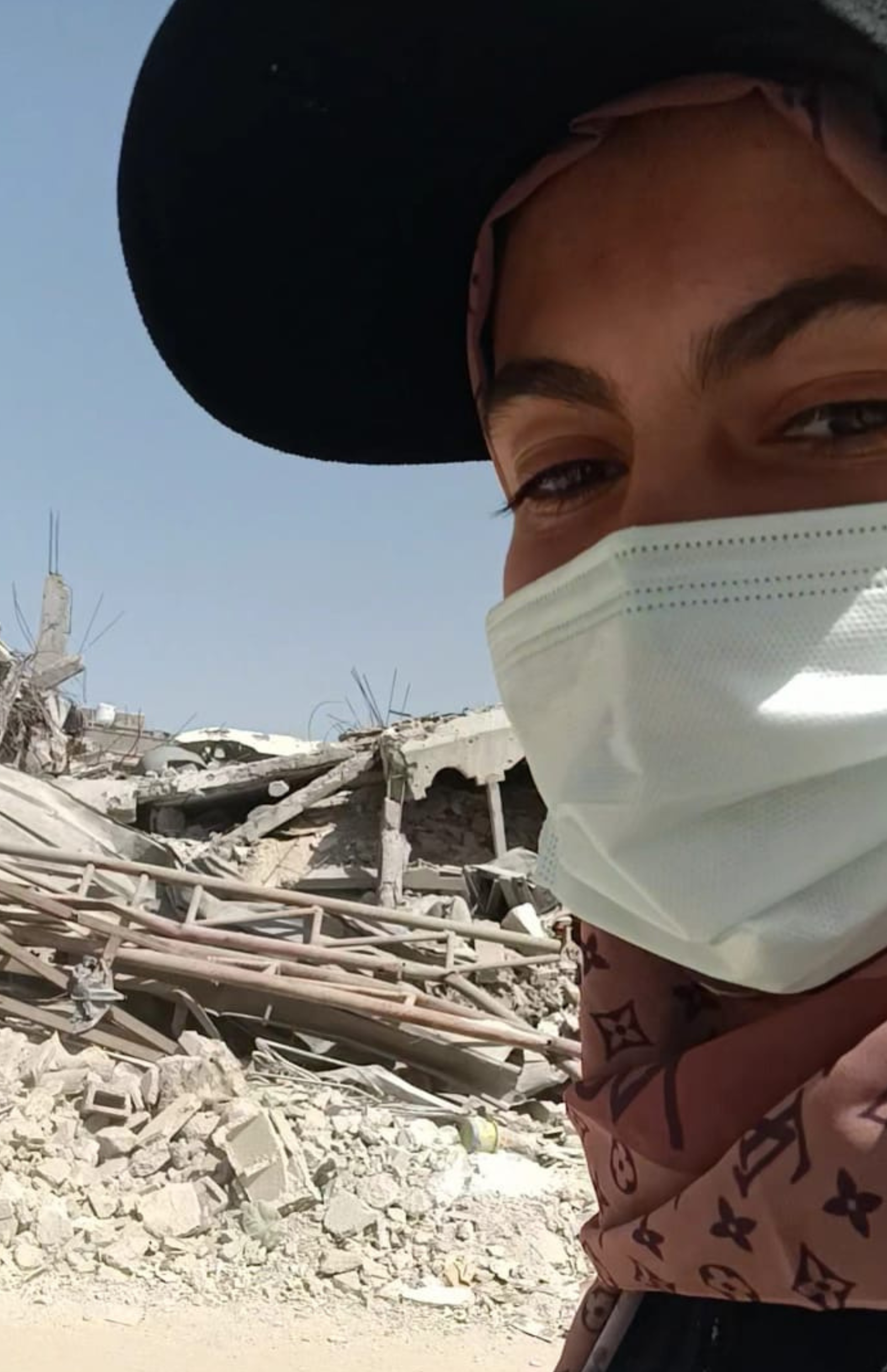 A person wearing a face mask and a large black hat standing amidst debris and destruction from a collapsed building or earthquake damage.