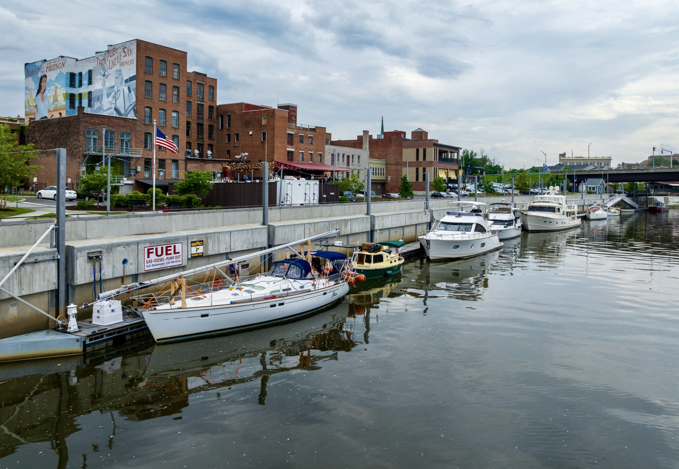 A marina with several docked boats and yachts along a concrete wall, with a cityscape featuring brick buildings, trees, and a cloudy sky in the background.