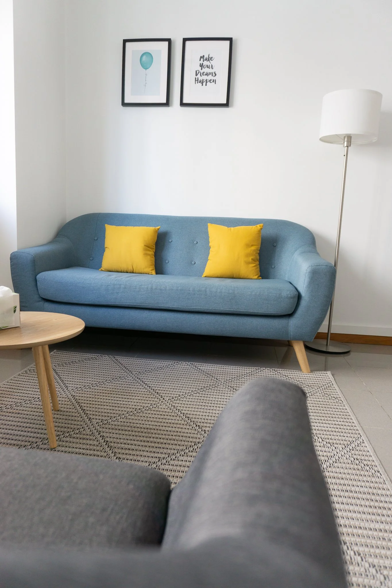 Sala de estar com sofá azul, duas almofadas amarelas, quadro na parede com um balão azul e uma frase, e uma luminária de pé.