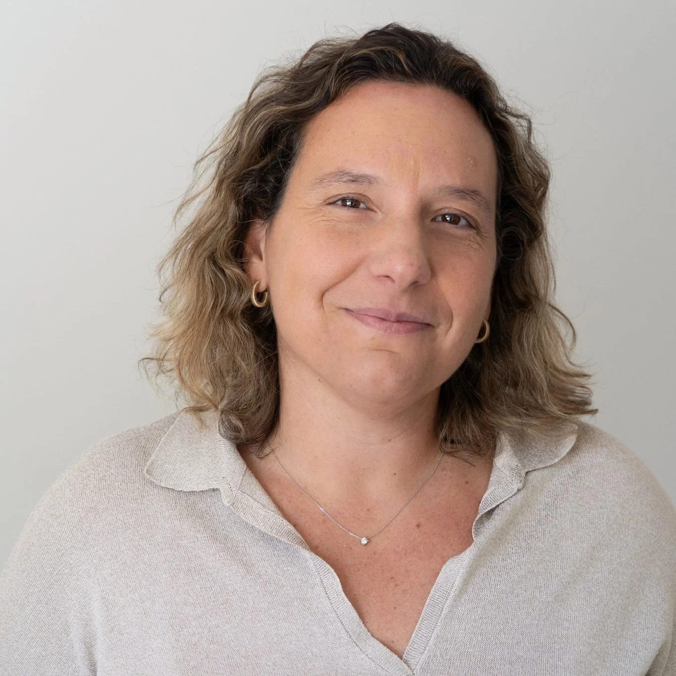 Retrato de mulher com cabelos cacheados, sorrindo, usando camisa bege e joias, contra fundo branco.