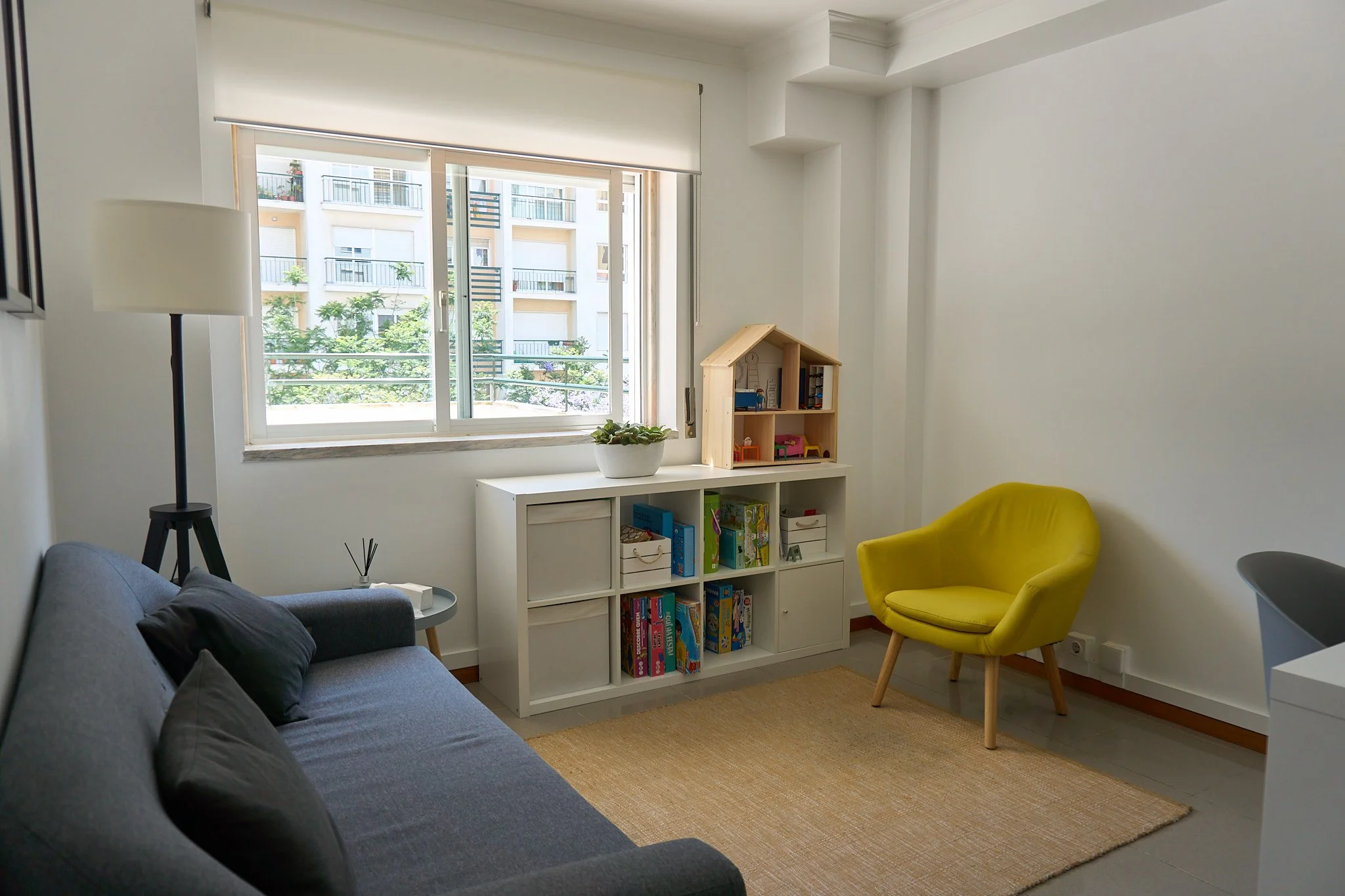 Sala de estar com sofá cinza, poltrona amarela, estante branca com brinquedos e livros, janela grande com vista para edifícios, tapete de fibra, luminária de pé preta.