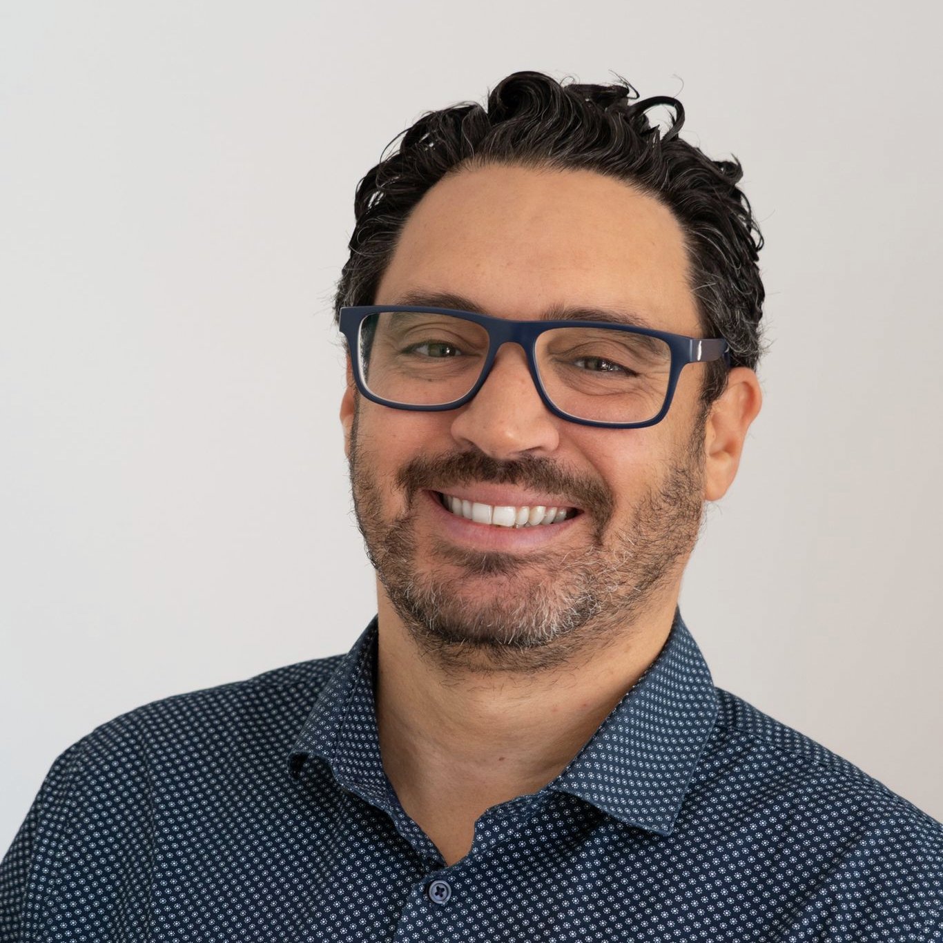 Homem de cabelo escuro e barba, usando óculos e camisa de manga longa com fundo branco.