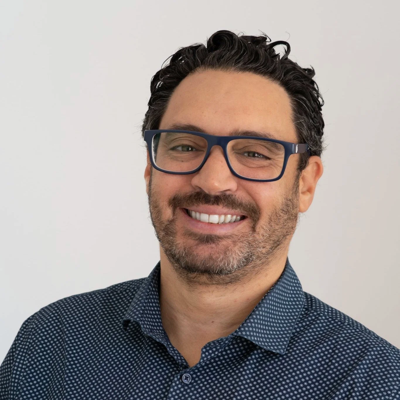 Homem sorridente de óculos escuros e barba, usando camisa de botão, contra fundo branco.