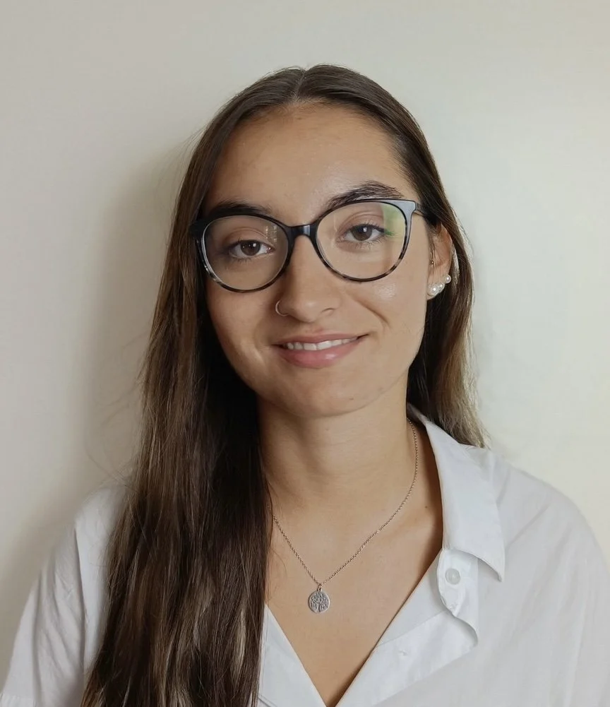 Retrato de uma jovem mulher com cabelos longos, óculos de armação preta, brincos de pérola, sorrindo, usando camisa branca e um colar, contra fundo branco