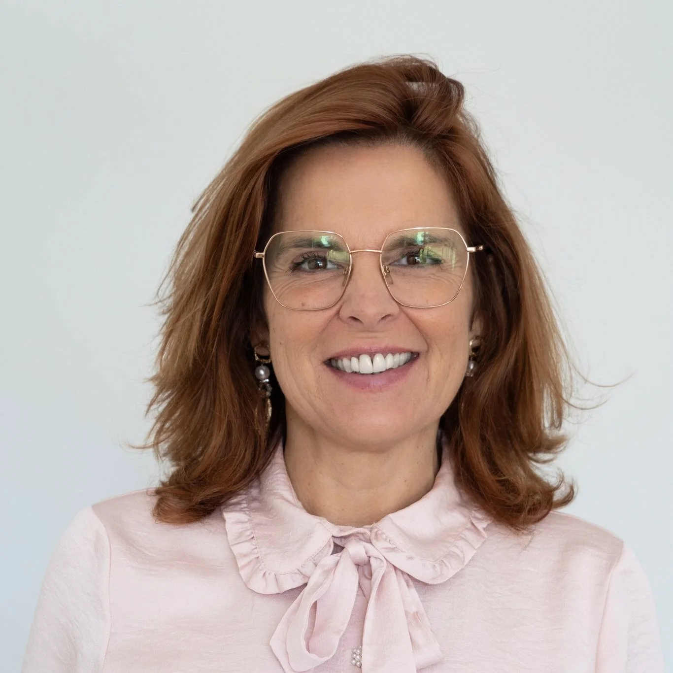 Mulher sorridente de cabelo castanho com óculos grandes de armação dourada, usando blusa rosa com laço no colarinho, contra fundo branco.