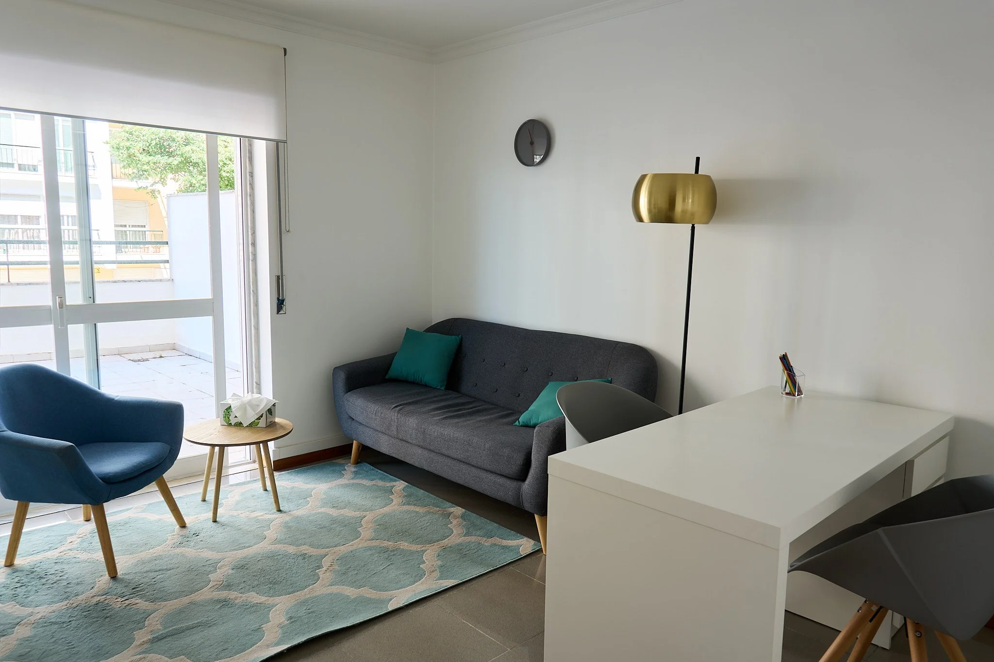 Sala de estar com sofá cinza, duas almofadas verdes, armário branco, cadeira preta, luminária dourada, tapete azul claro com padrão branco, e varanda com porta de vidro.