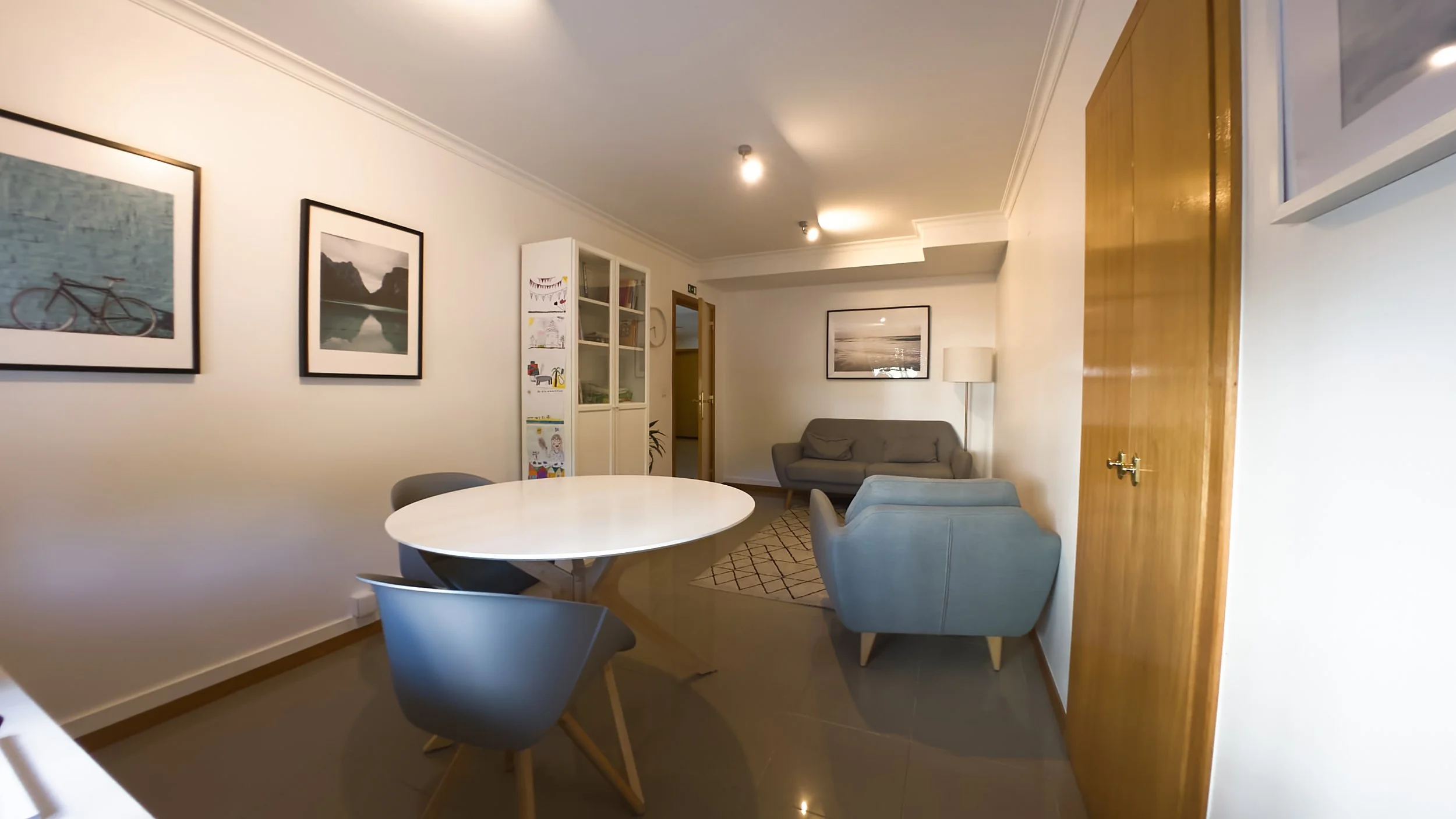 Sala de estar com mesa redonda, cadeiras cinzas, sofá cinza, detalhes em branco, quadro na parede, luminária de pé e porta de madeira.