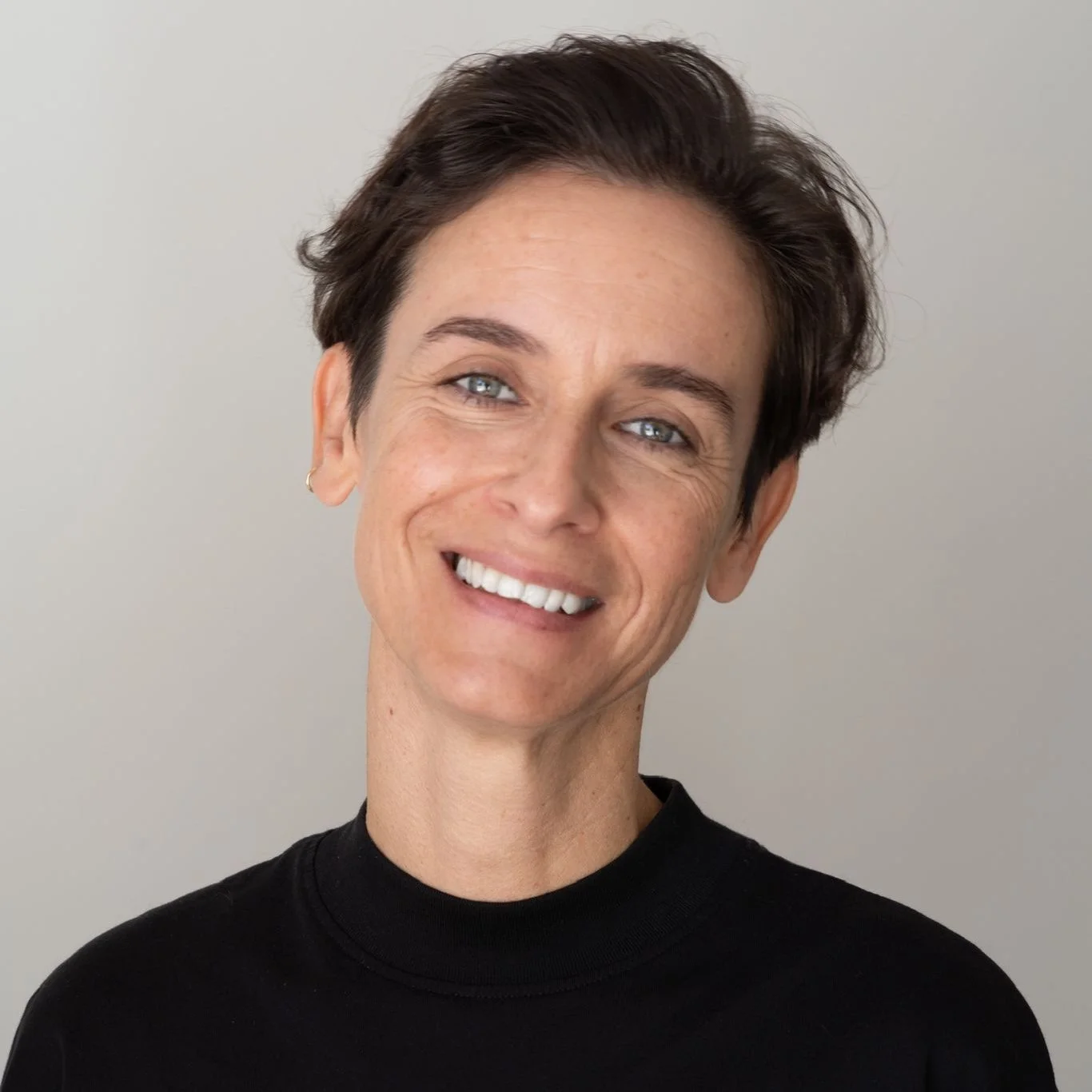 Retrato de uma mulher com cabelo curto, escuro, usando blusa preta, sorrindo, com fundo neutro.