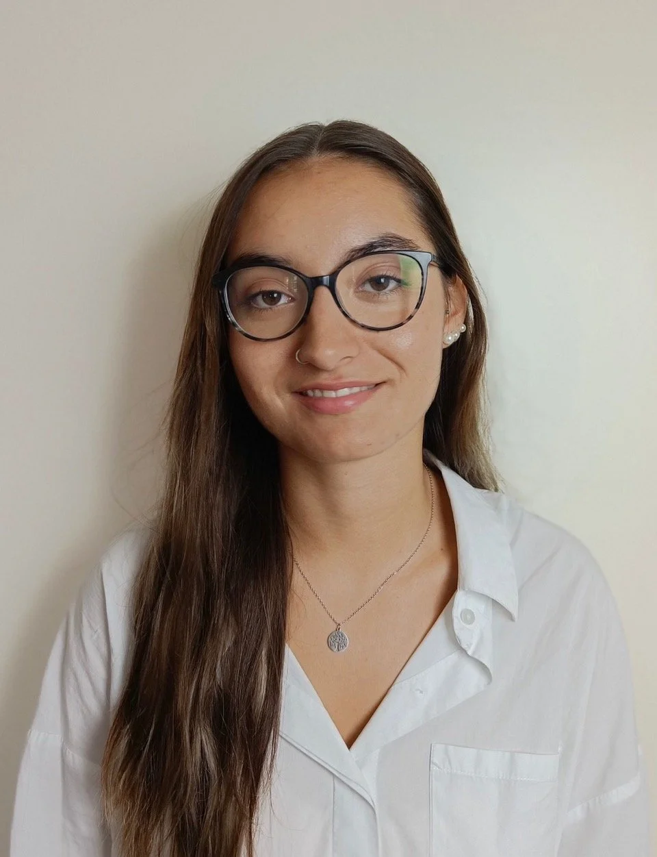 Retrato de uma jovem mulher com cabelos longos e castanhos, óculos de armação preta, brincos de pérola, usando camisa branca e colar com pingente de árvore, sorrindo levemente na frente de uma parede clara.