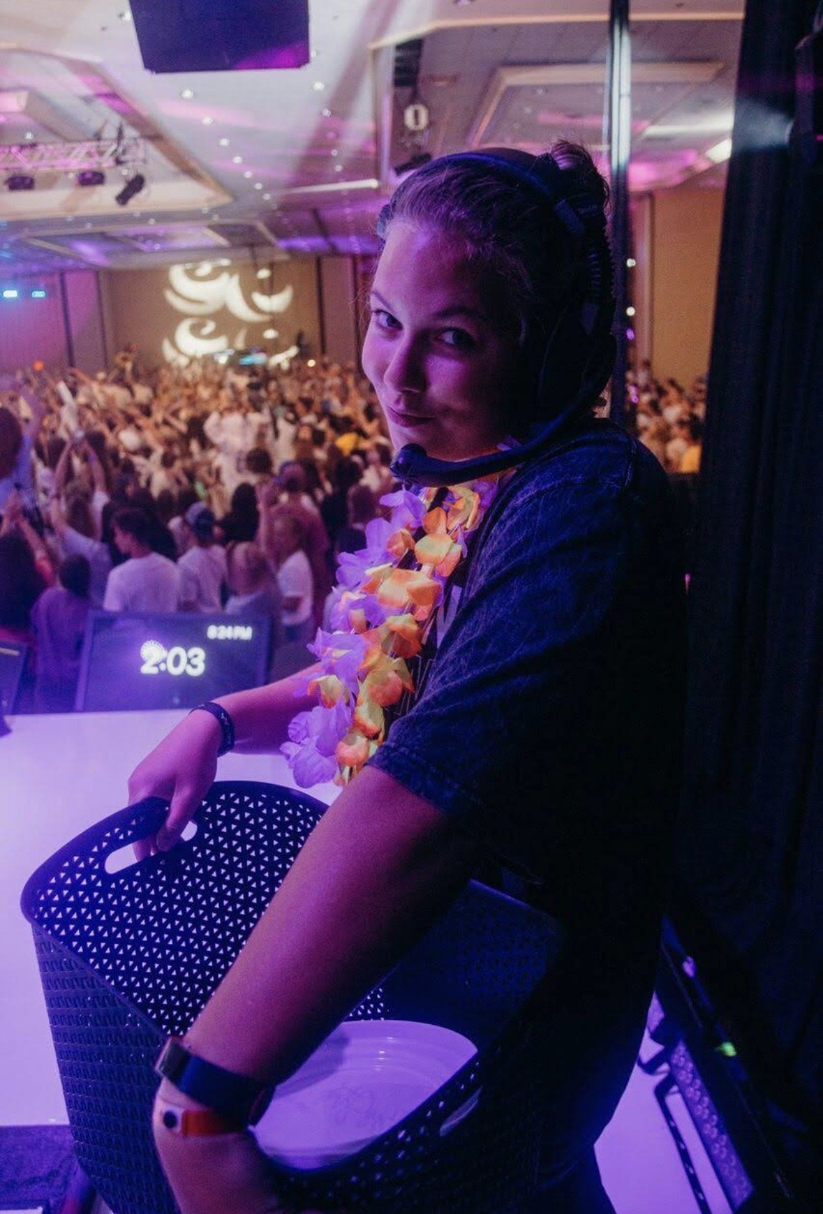 A person wearing a floral lei, headphones, and a dark shirt, standing behind a DJ booth at a crowded indoor event with colorful lights and people dancing in the background.