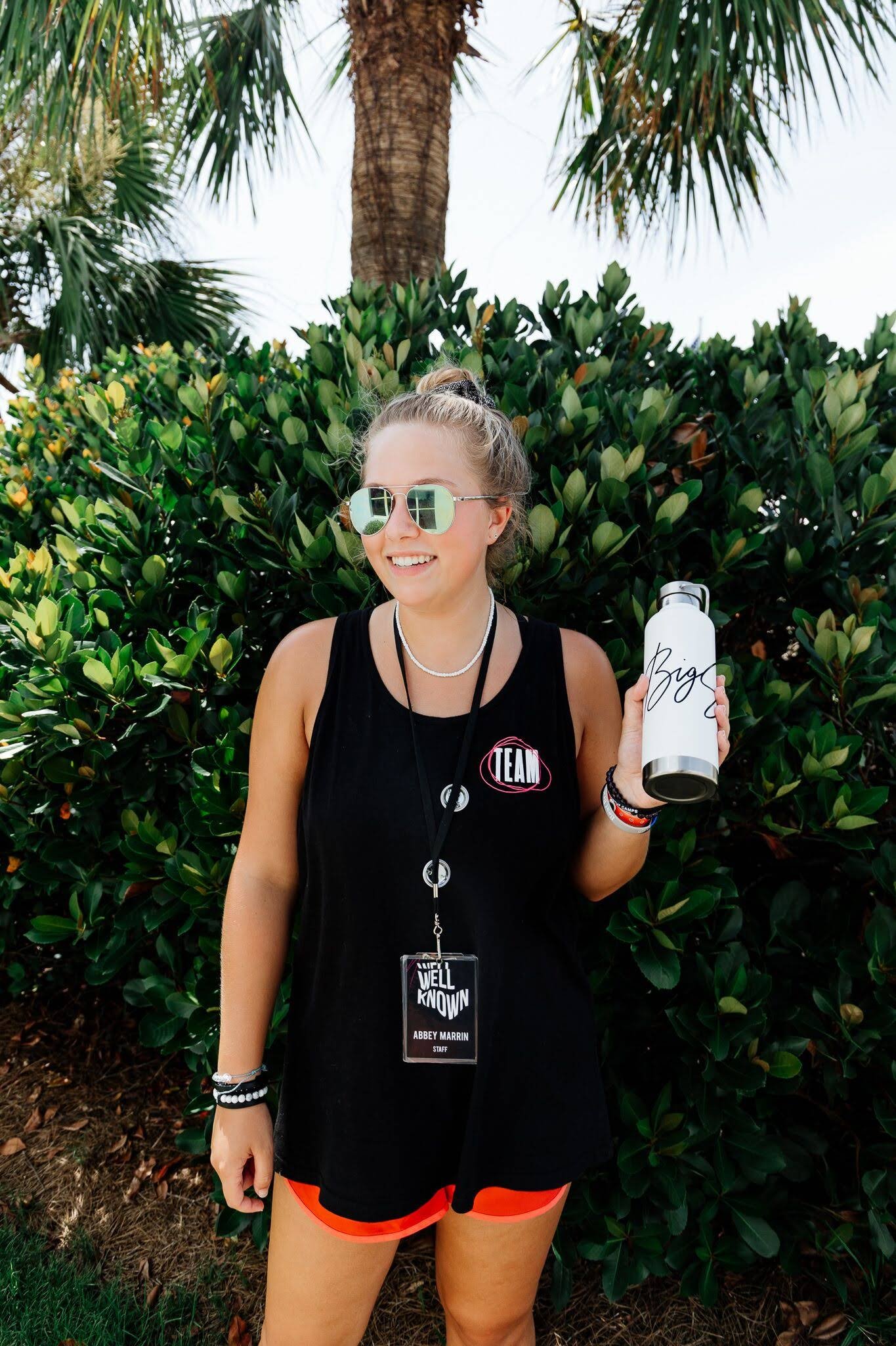 A smiling young woman with sunglasses, wearing a black sports tank top and orange shorts, holding a white water bottle with 'Big' written on it, standing in front of green foliage and a palm tree.