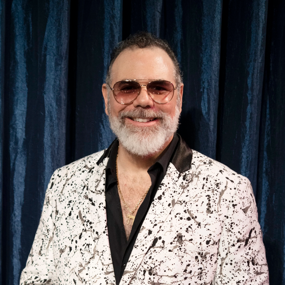 A man with a white beard and glasses smiling, wearing a black shirt and a white blazer with black abstract splatters, standing in front of a dark blue curtain. Adam Realman - Coney Island Circus Sideshow Artistic Director