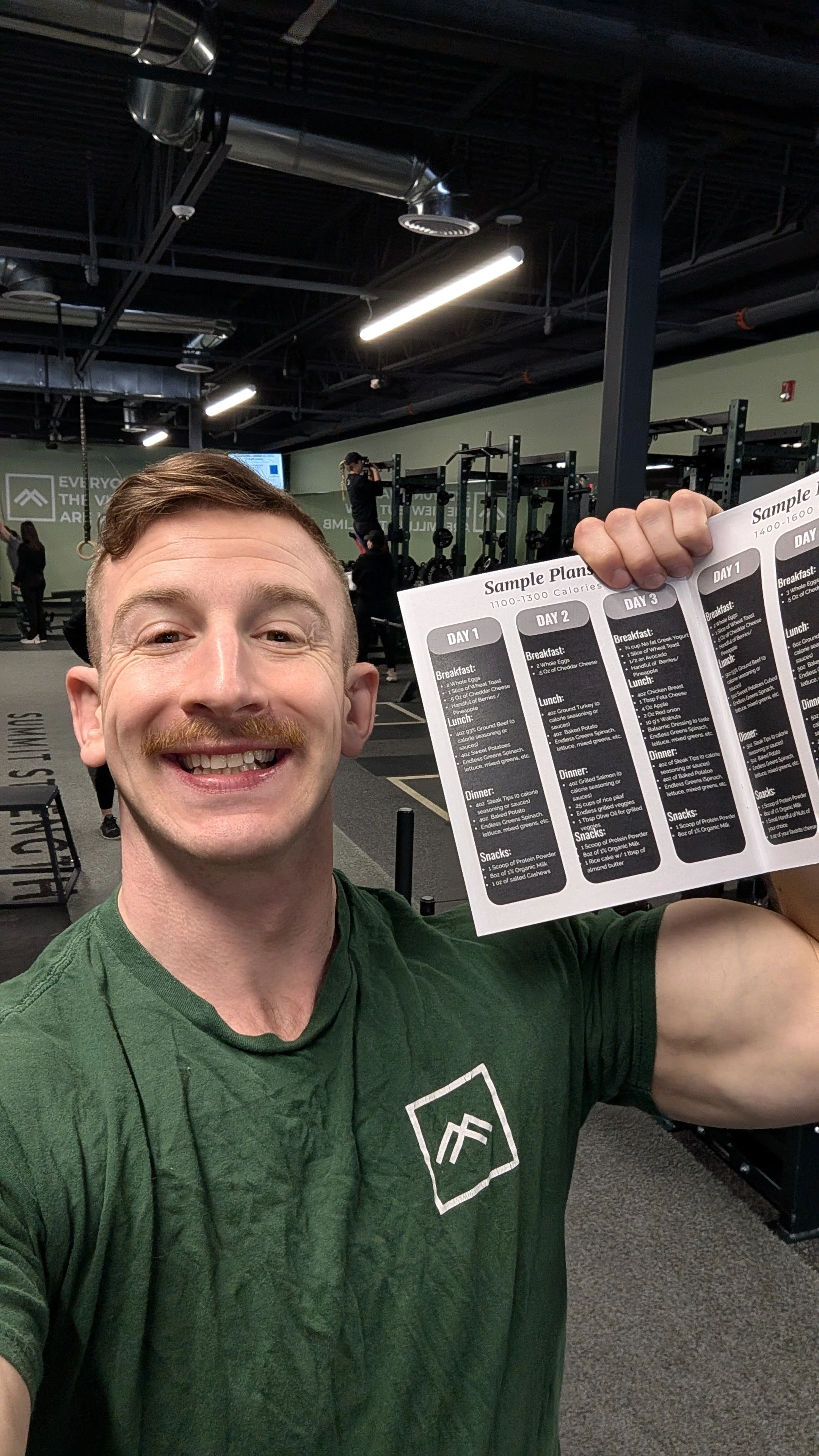 A smiling man in a green shirt holding a sample meal plan at the gym, with gym equipment and other people working out in the background.