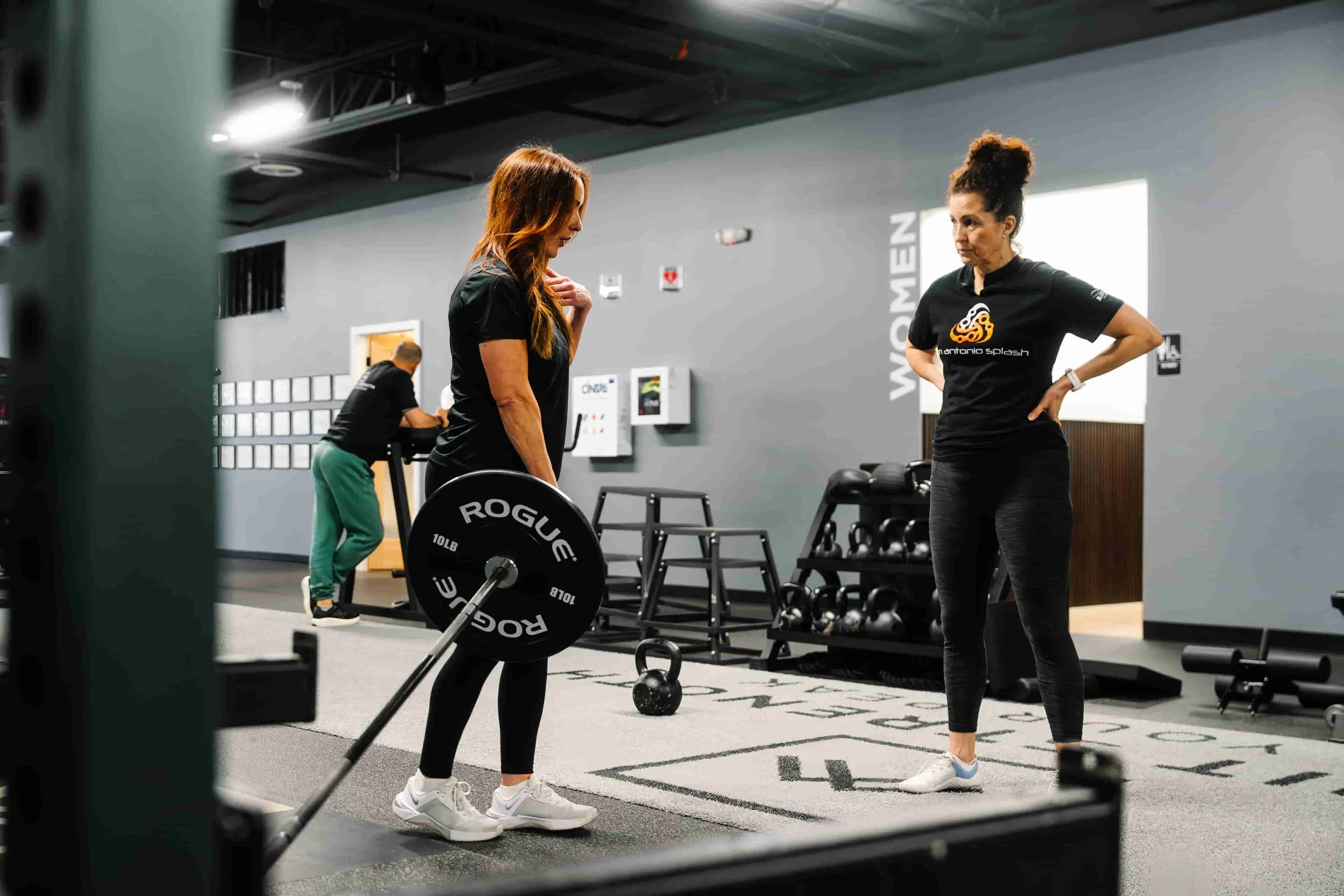 A female personal trainer attentively coaching a woman performing a barbell deadlift with Rogue weights at a private training gym in Milford, New Hampshire