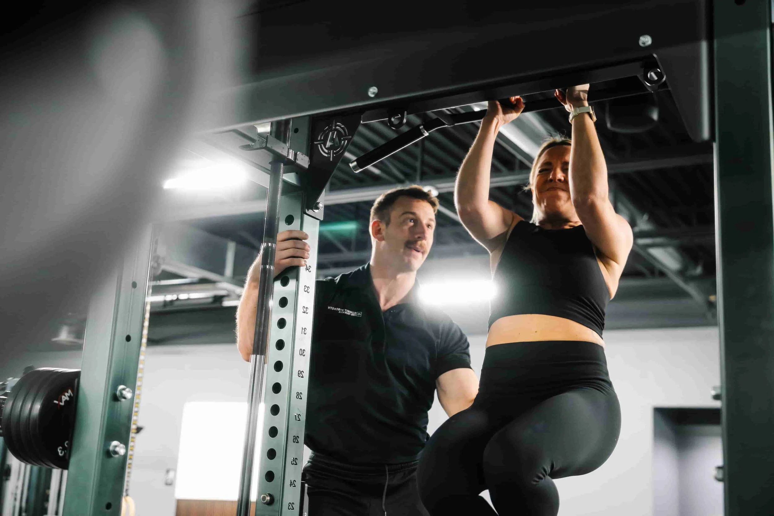 A male personal trainer spotting and encouraging a female client performing a pull-up on a power rack at a private strength training gym in Milford, New Hampshire