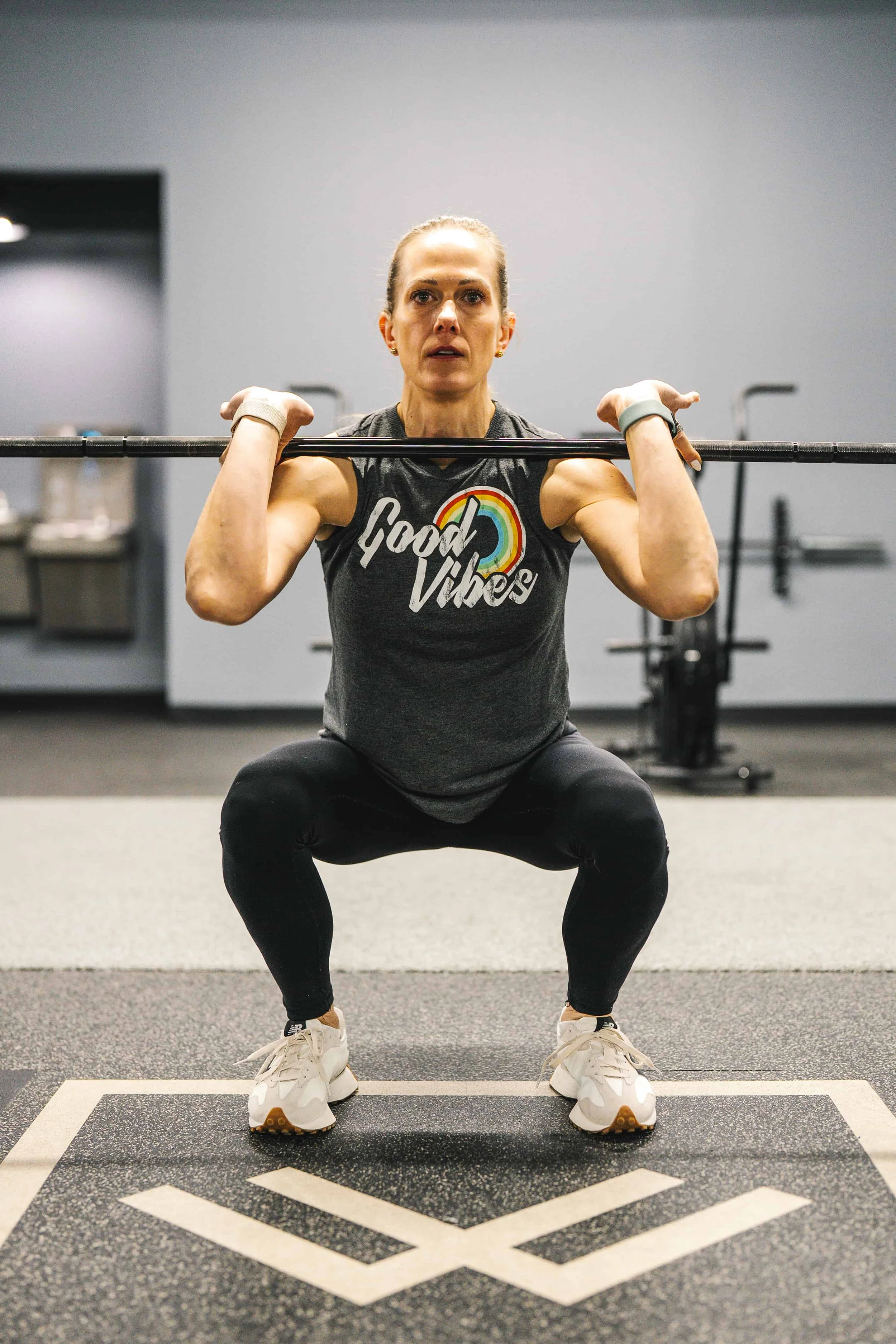 A woman wearing a sleeveless shirt with 'Good Vibes' and a rainbow print, squatting with a barbell on her shoulders in a gym.