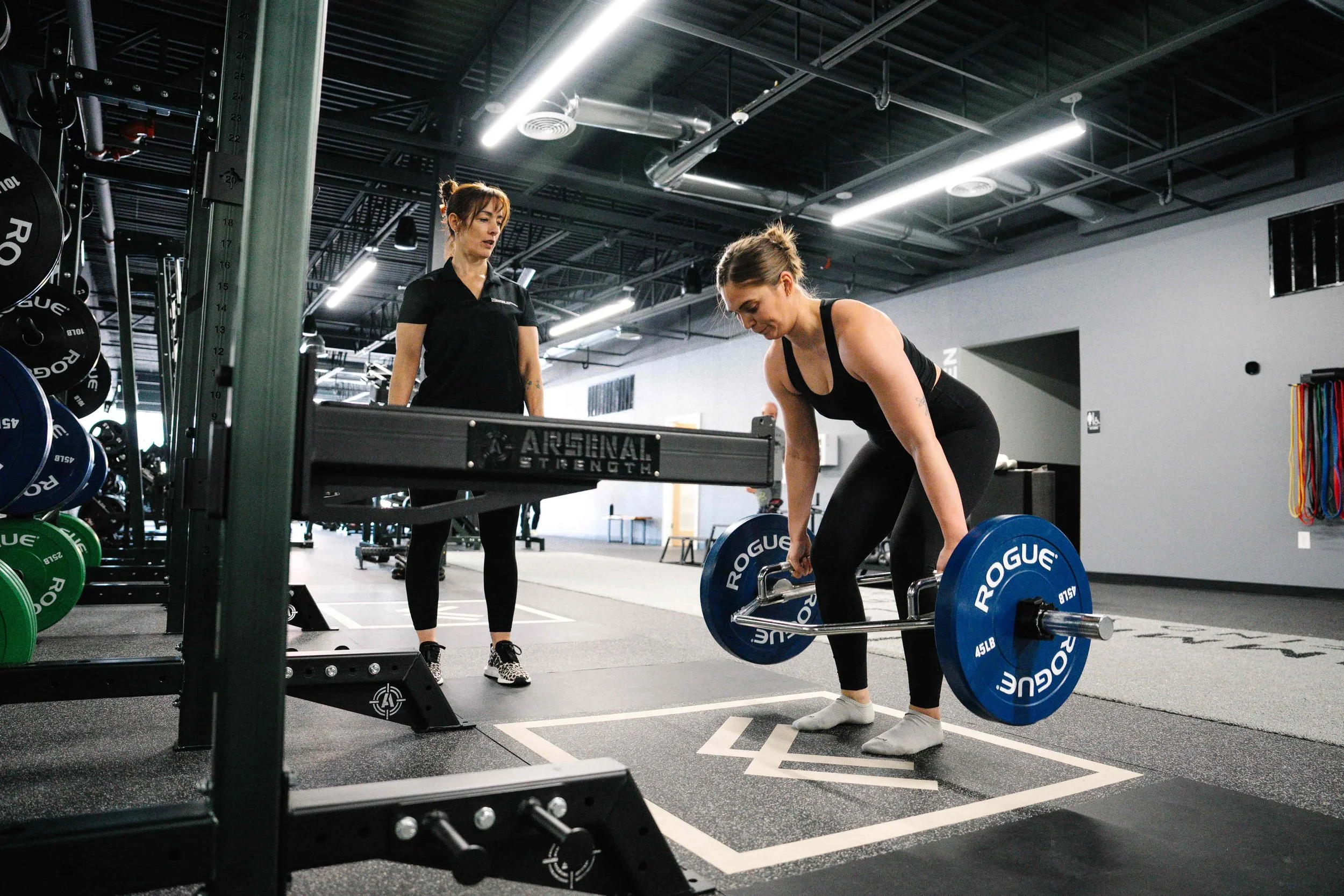 A woman in black athletic wear is lifting a blue Rogue barbell with weights in a gym. A coach or trainer observes nearby, standing next to a weightlifting platform in a fitness facility.