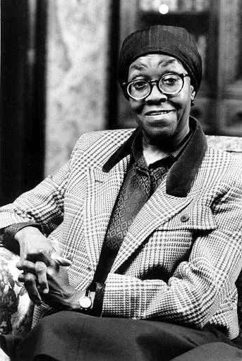 Gwendolyn Brooks in a plaid coat and black turban. An African American woman with round glasses holding her hands intertwined over the arm of a chair.