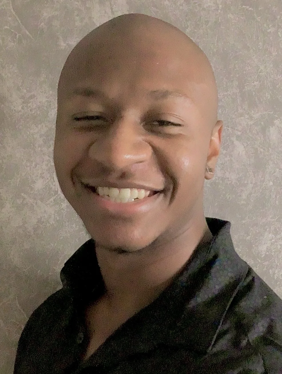 A smiling man with a shaved head, light skin, and earrings, wearing a black shirt, standing against a textured gray wall.