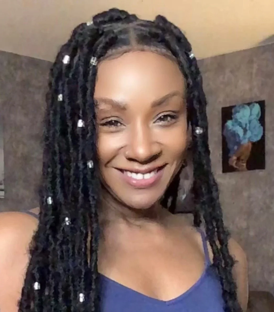 A woman smiling with black dreadlocks decorated with white beads, wearing a purple top, standing in a room with textured walls and framed artwork.