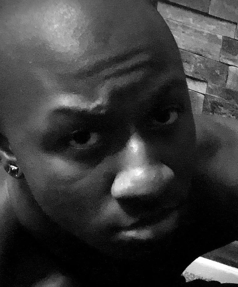Black and white close-up photo of a person with a shaved head, dark eye makeup, and an earring, looking directly at the camera.