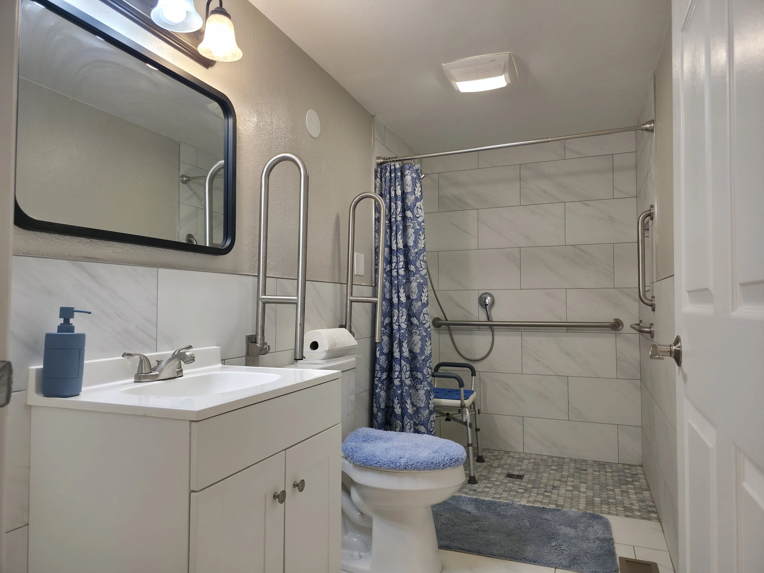 Accessible bathroom with white sink and cabinet, mirror, toilet with blue seat cover, towel, walk-in shower with grab bars, shower chair, and blue patterned shower curtain.