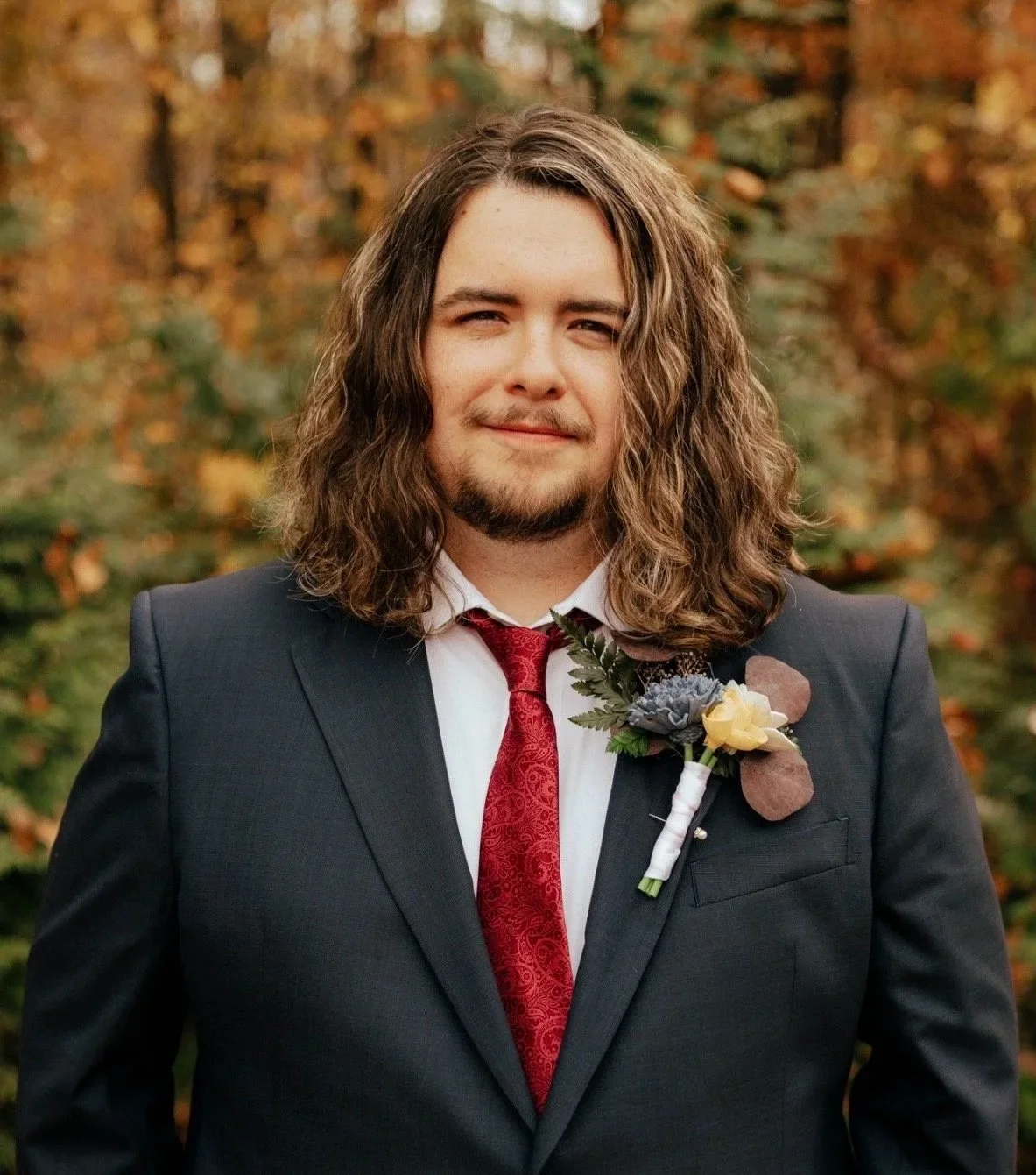 A man dressed in a black suit with a red tie, wearing a boutonniere, standing outdoors with fall foliage in the background.